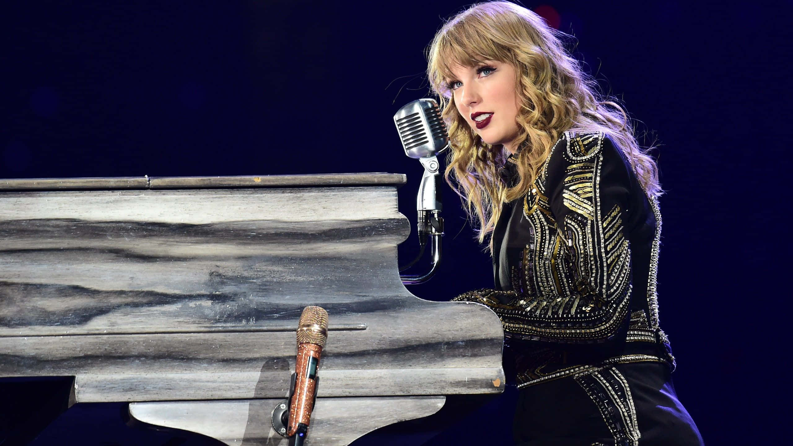 泰勒·斯威夫特《Folklore》世界巡迴演唱會 壁紙 — Taylor Swift 在舞台燈光下，用復古咪高峰在鋼琴前熱情演出。