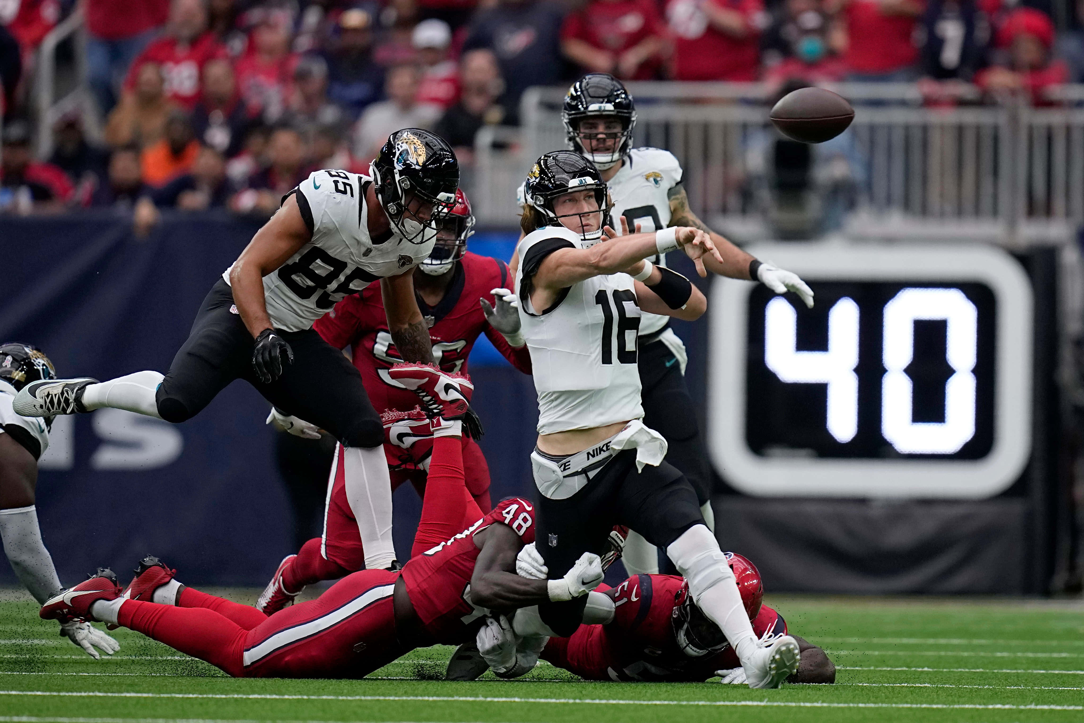 Trevor Lawrence 桌布 壁紙 — 一名身穿白色球衣的美式足球四分衛在綠色球場上被紅色防守球員擒抱時擲球。