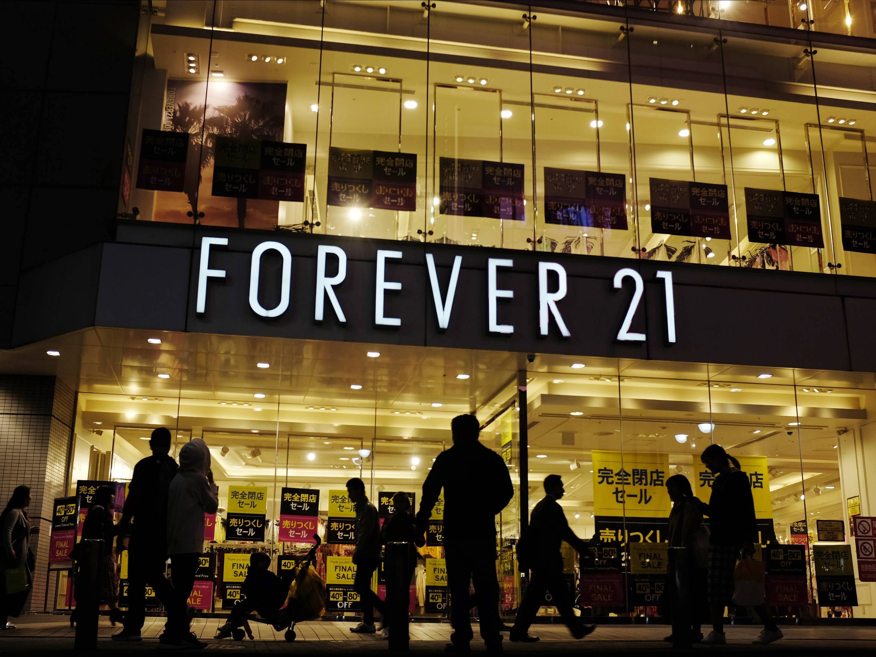 Forever 21日本分店 壁紙 —  silhouetted 嘅行人夜晚行過一間燈火通明嘅 Forever 21 舖頭，玻璃窗入面見到日本嘅減價標誌。