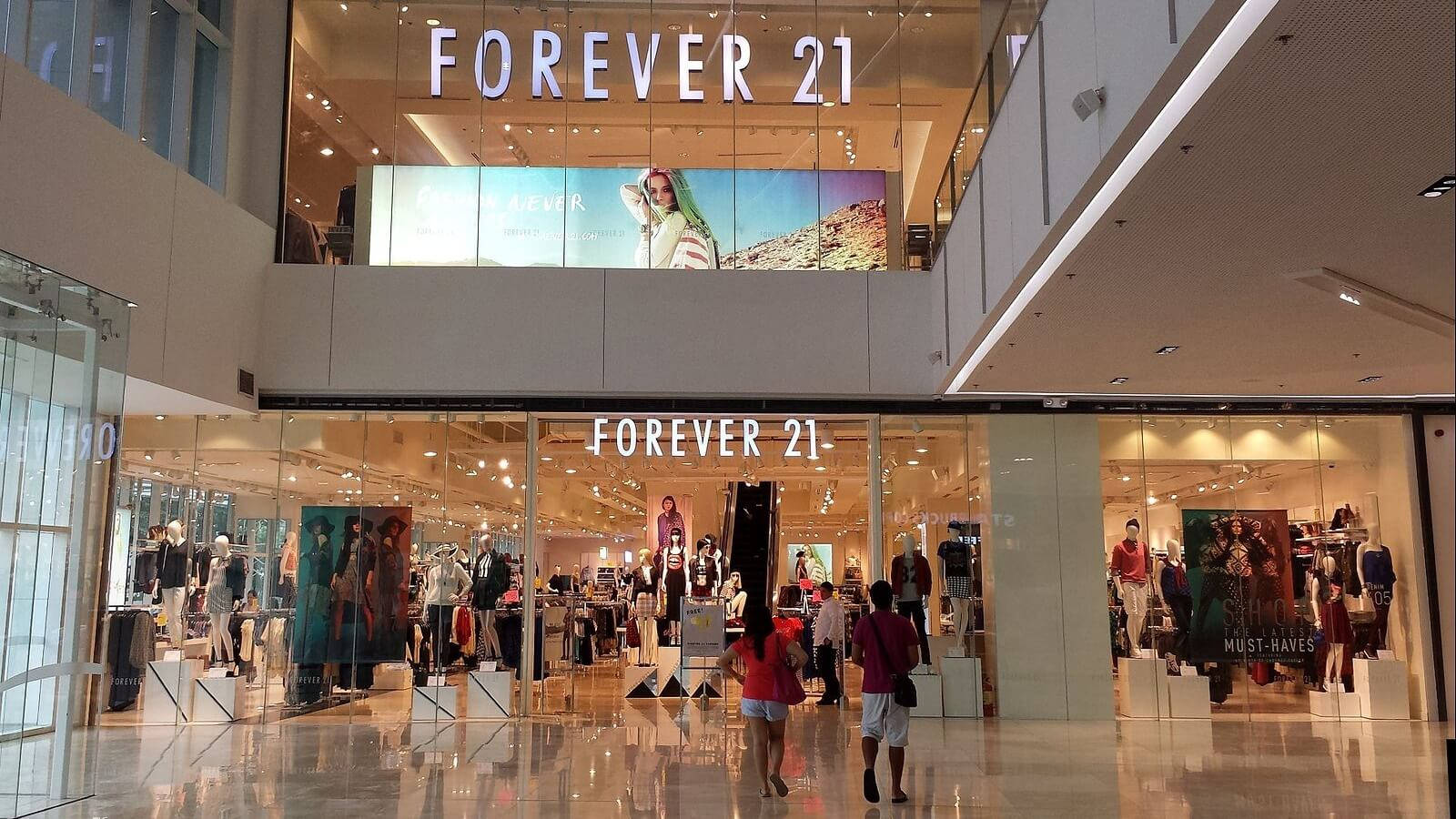 Forever 21購物中心內景 壁紙 — 購物者走過一間燈光明亮的Forever 21商店，位於一個現代化購物中心內，地板反光，並有大型玻璃窗。