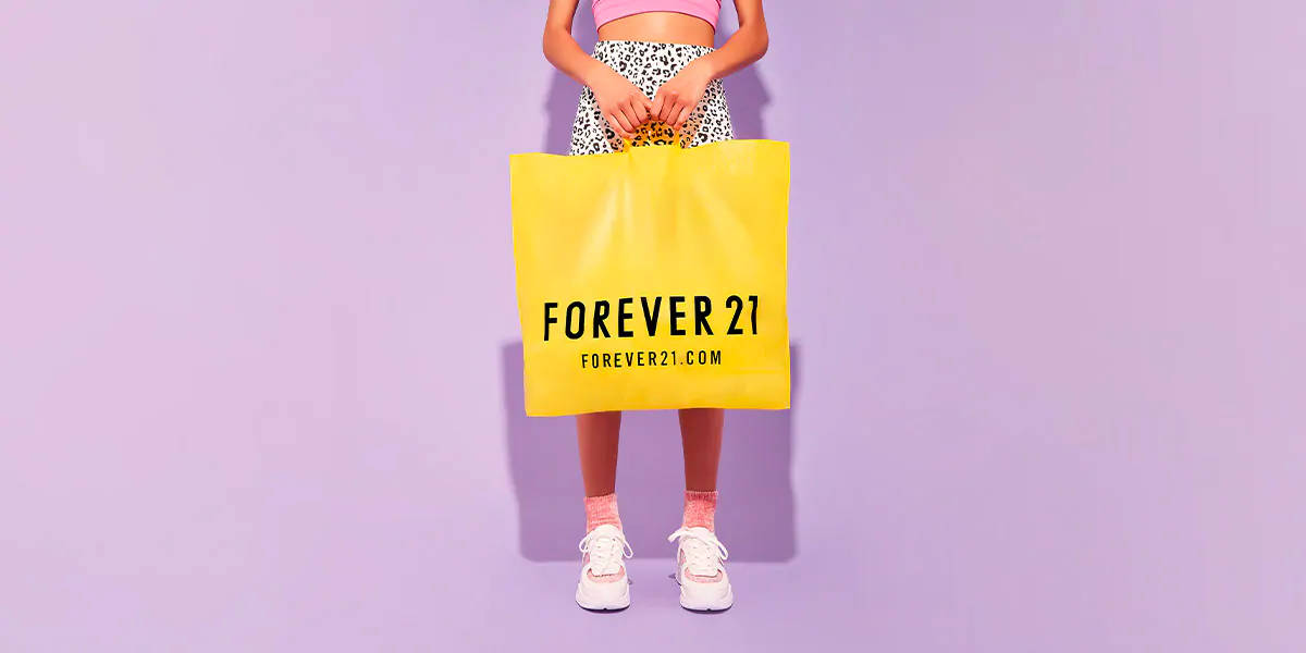 Forever 21黃色購物袋 壁紙 — 一位身穿粉紅色和豹紋圖案的人，在純色薰衣草背景前拿著一個亮黃色的Forever 21購物袋。