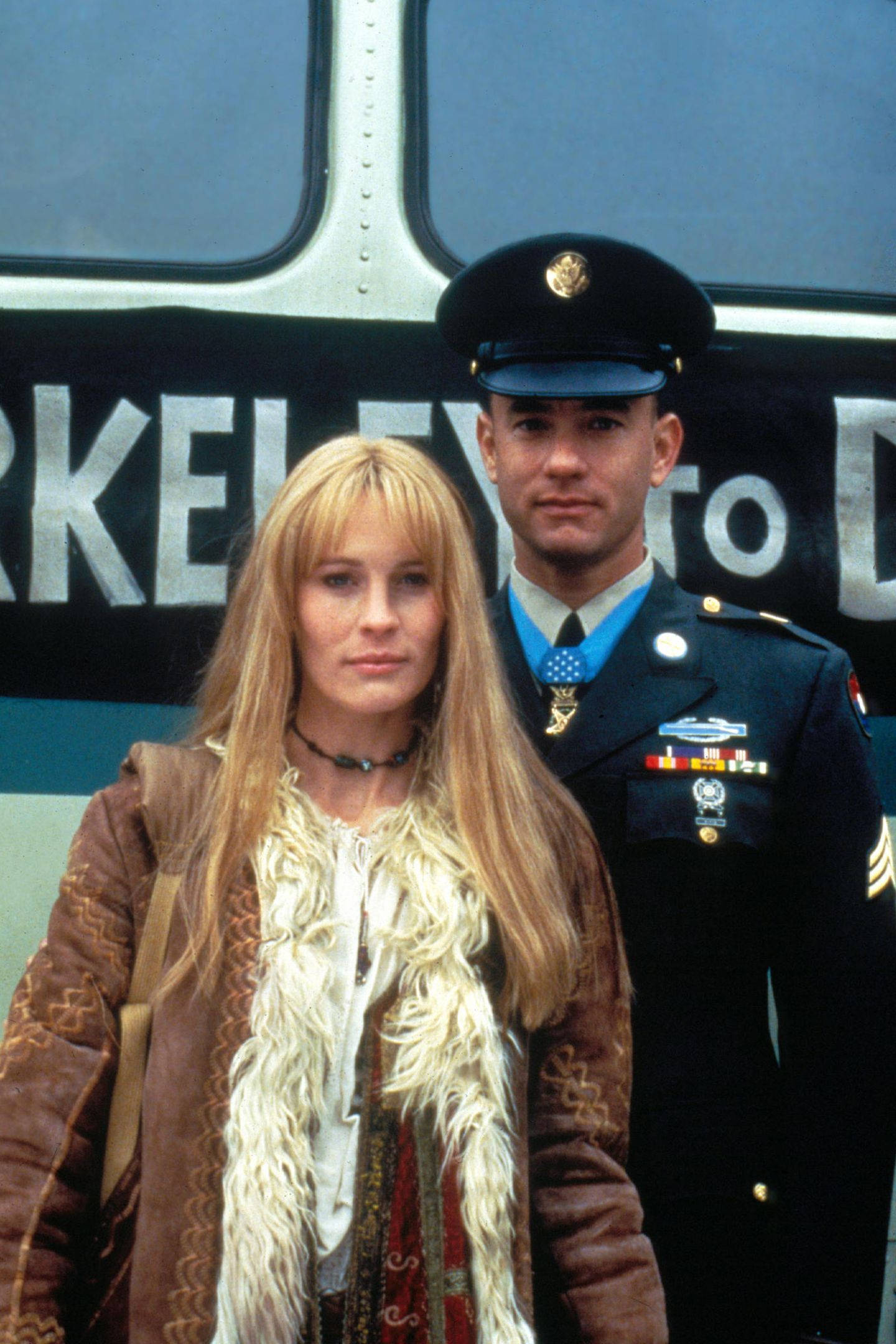 Forrest Gump 與 Jenny 站在一起 壁紙 — 一位穿著流蘇棕色外套的女性站在一輛巴士前，旁邊是一位穿著制服、佩戴勳章的士兵。