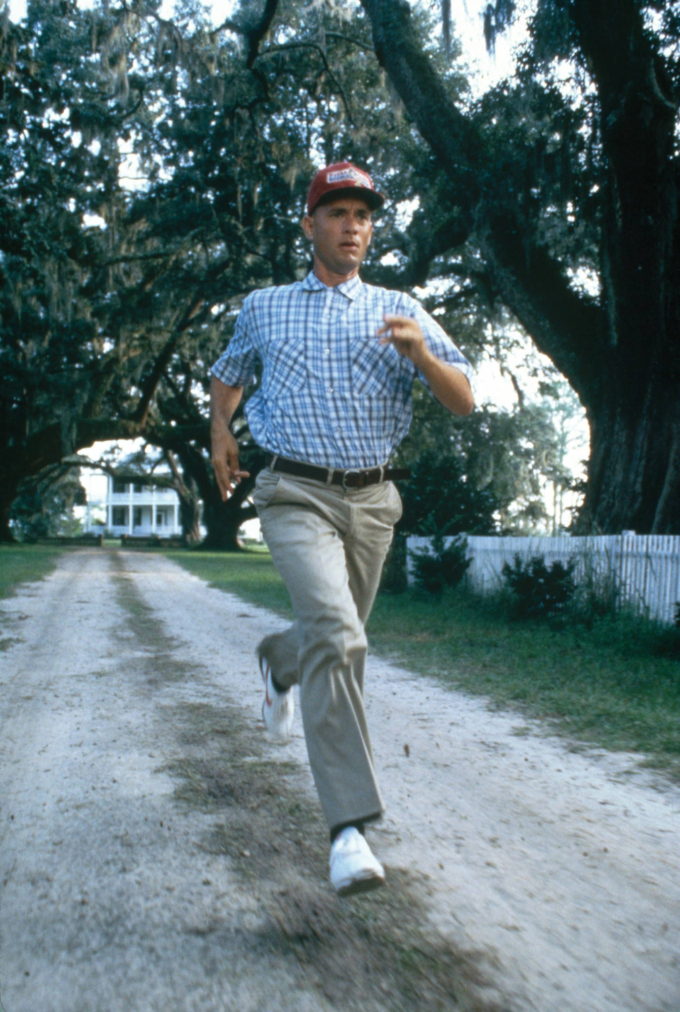 Forrest Gump 在土路上奔跑 壁紙 — 一名身穿格仔衫、戴紅色帽嘅男人喺兩旁有樹嘅泥路上向住一間白色屋跑。