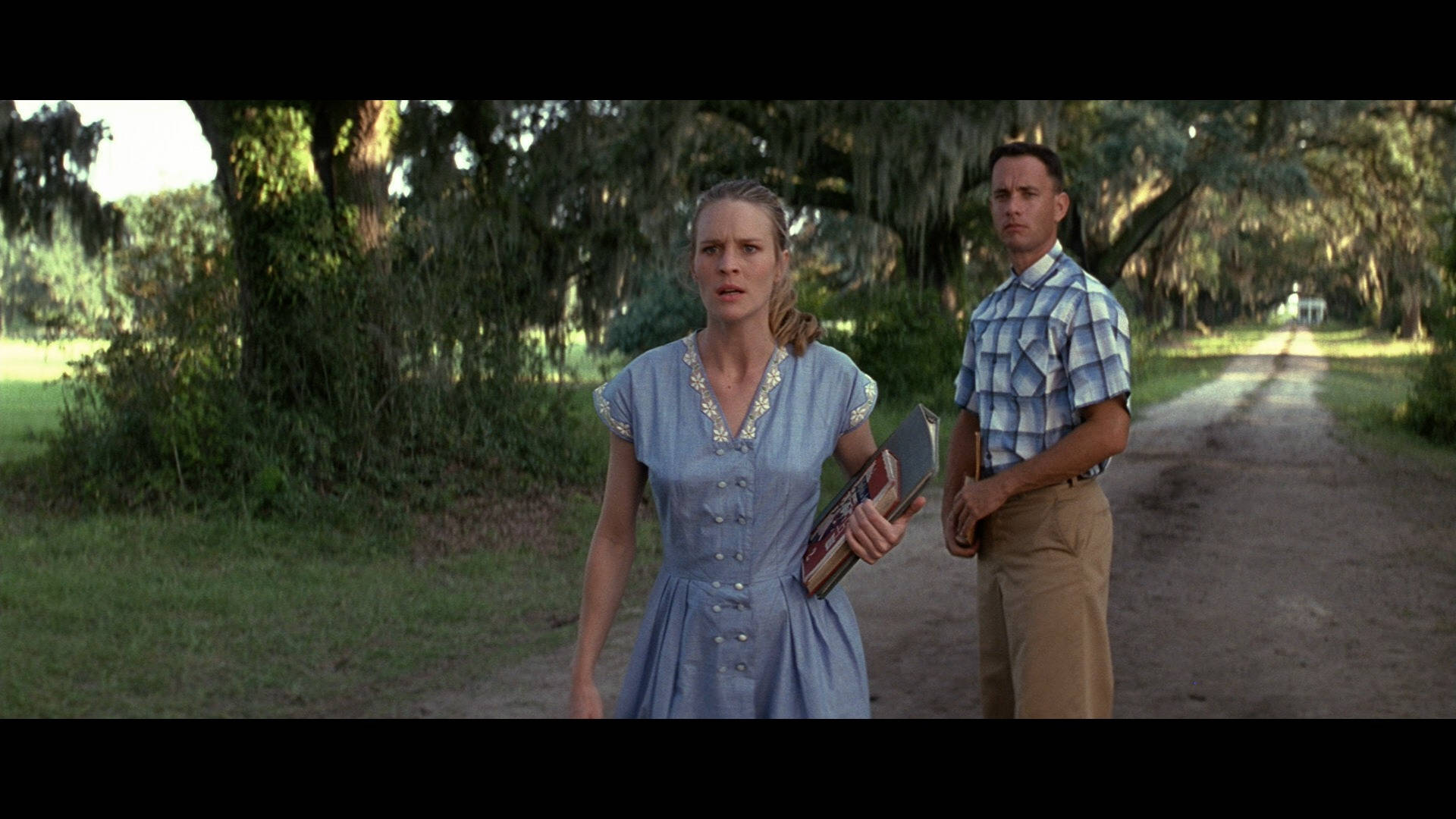 Forrest Gump 與 Jenny 身穿藍色 壁紙 — 一位身穿藍色連衣裙的女子和一位身穿格仔襯衫的男子站在一條陽光普照的泥路上，路旁長滿了掛滿苔蘚的樹木。