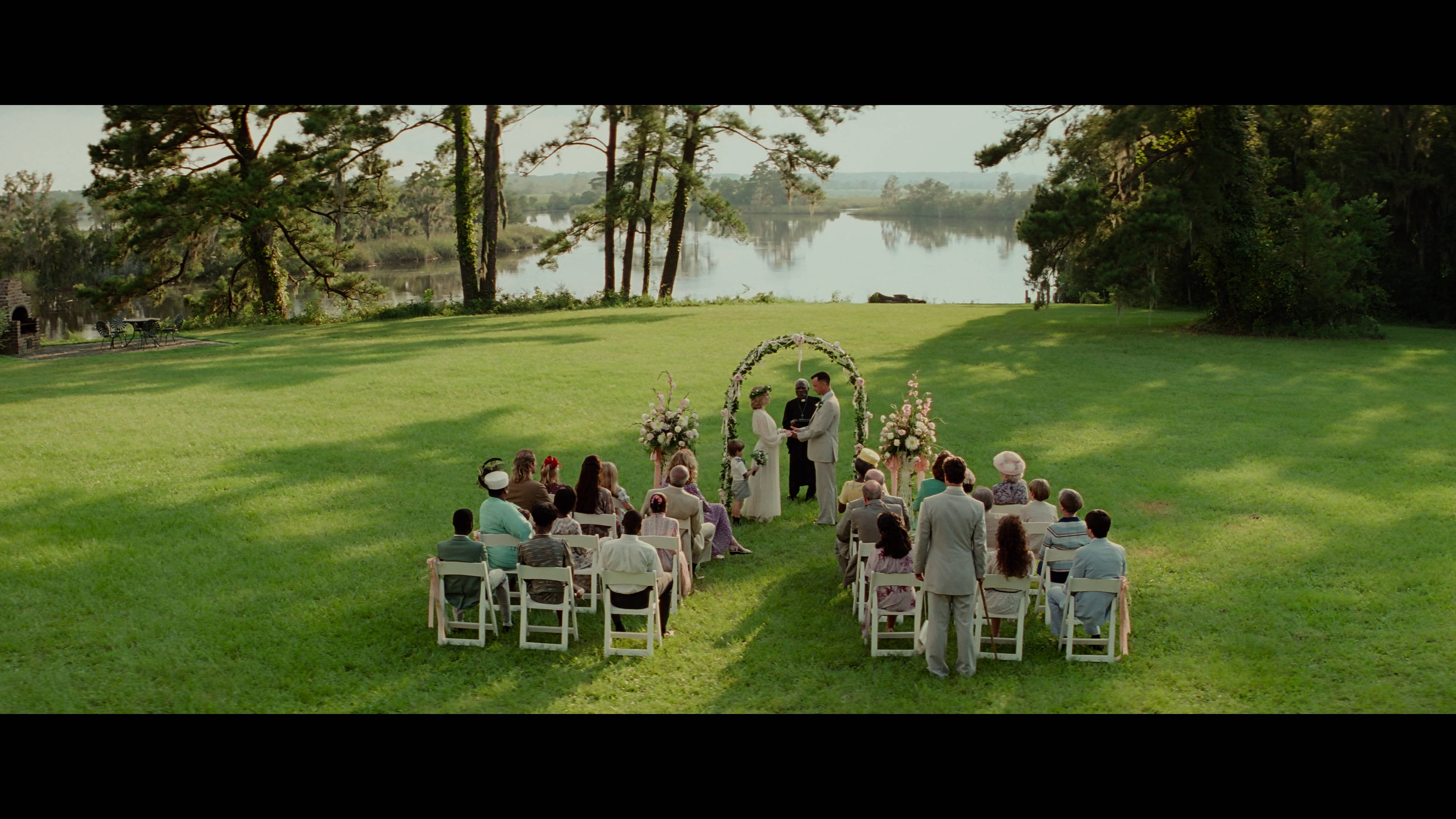 Forrest Gump 與 Jenny 婚禮 壁紙 — 一場婚禮在陽光明媚的草地上舉行，旁邊是平靜的湖泊，賓客坐在高樹下的白色椅子上。