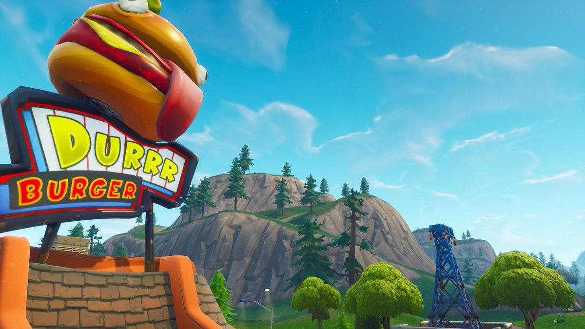 Fortnite 精選縮圖：Durr Burger 快餐店 Retail Row 場景 壁紙 — 一個巨大的卡通漢堡棲息在色彩繽紛的Durrr Burger招牌上，背景是明亮的藍天、岩石丘陵和樹木。