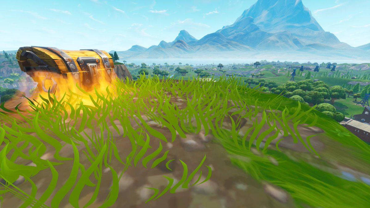 Fortnite 精選縮圖：草地與藍山腳下的戰鬥箱 壁紙 — 一個燃燒的寶箱放在翠綠的草地上，遠方有山脈，天空清澈湛藍。