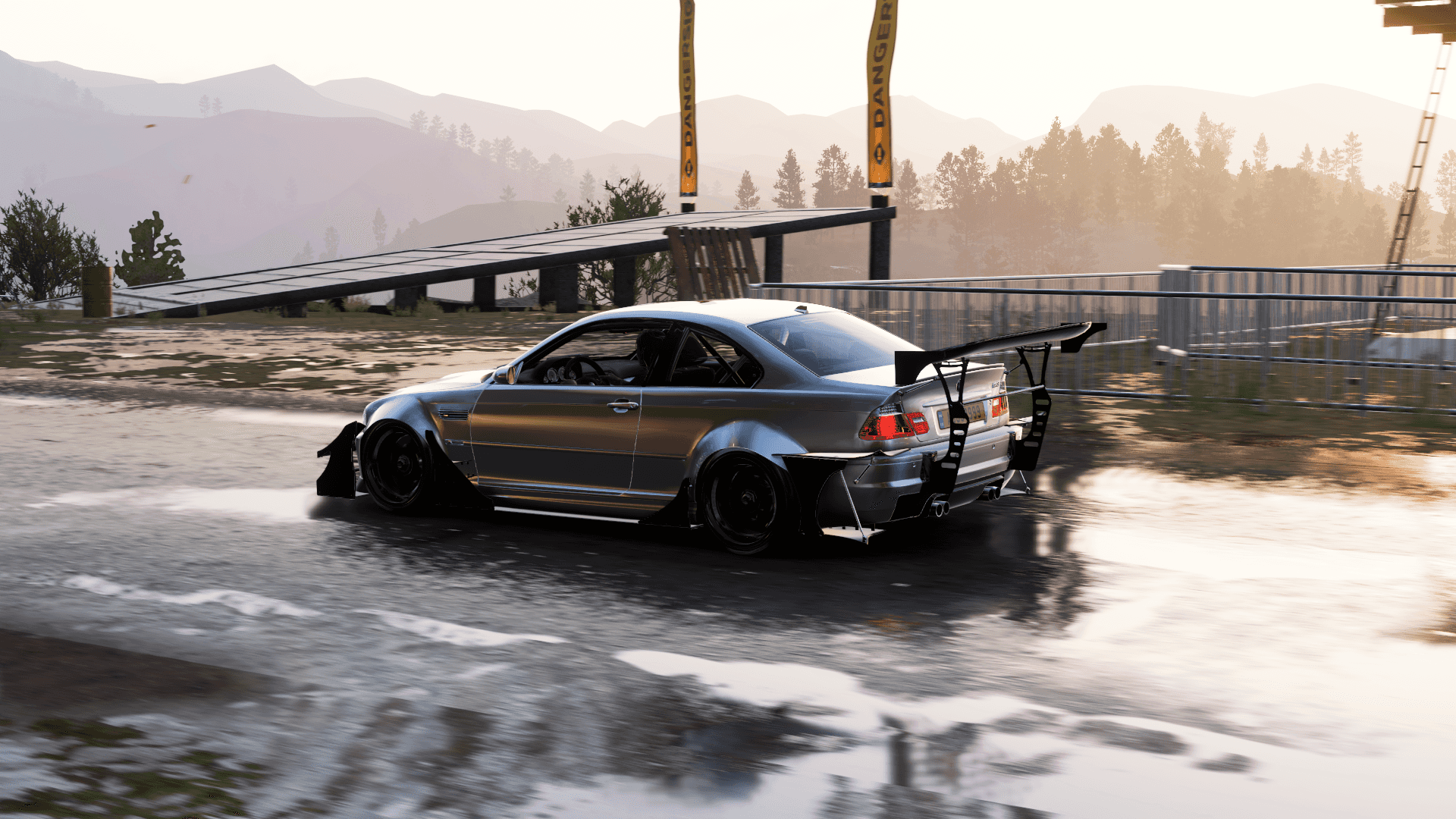 Forza Horizon - BMW 3系列 壁紙 — 一輛銀色飄移車帶有大型後尾翼，在濕滑的瀝青賽道上側滑，附近有橋樑，背景是山脈。