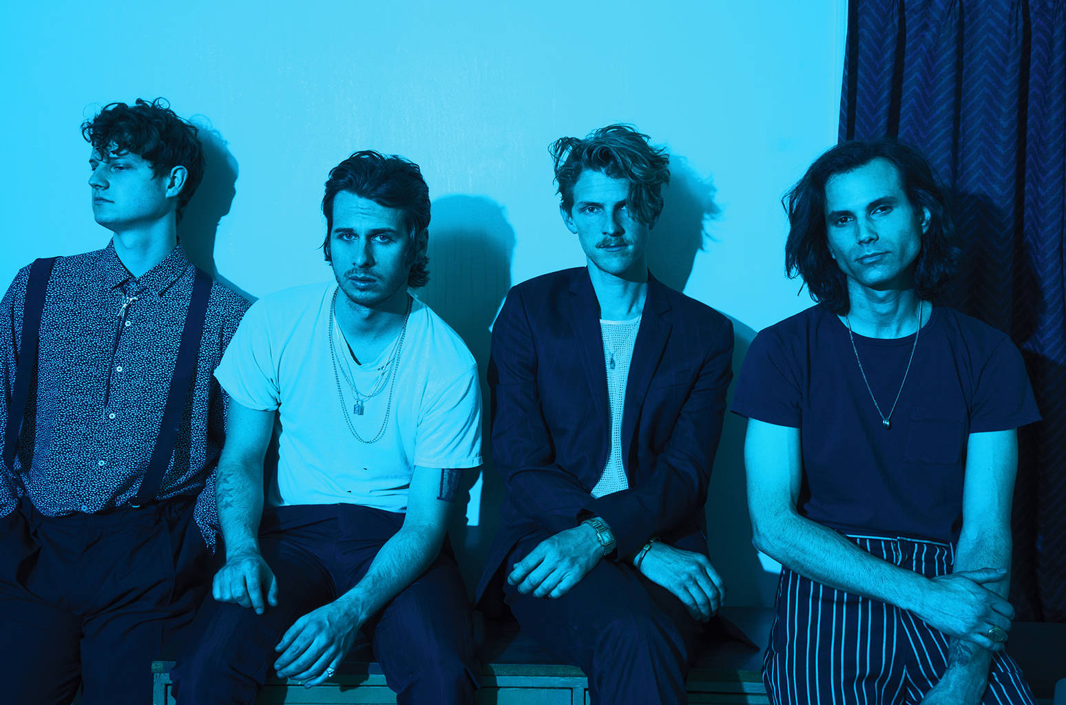 Foster The People 复古風貌 壁紙 — 四個男人排成一排，坐在藍色牆壁前，在冷色燈光下，各有不同的風格和表情。