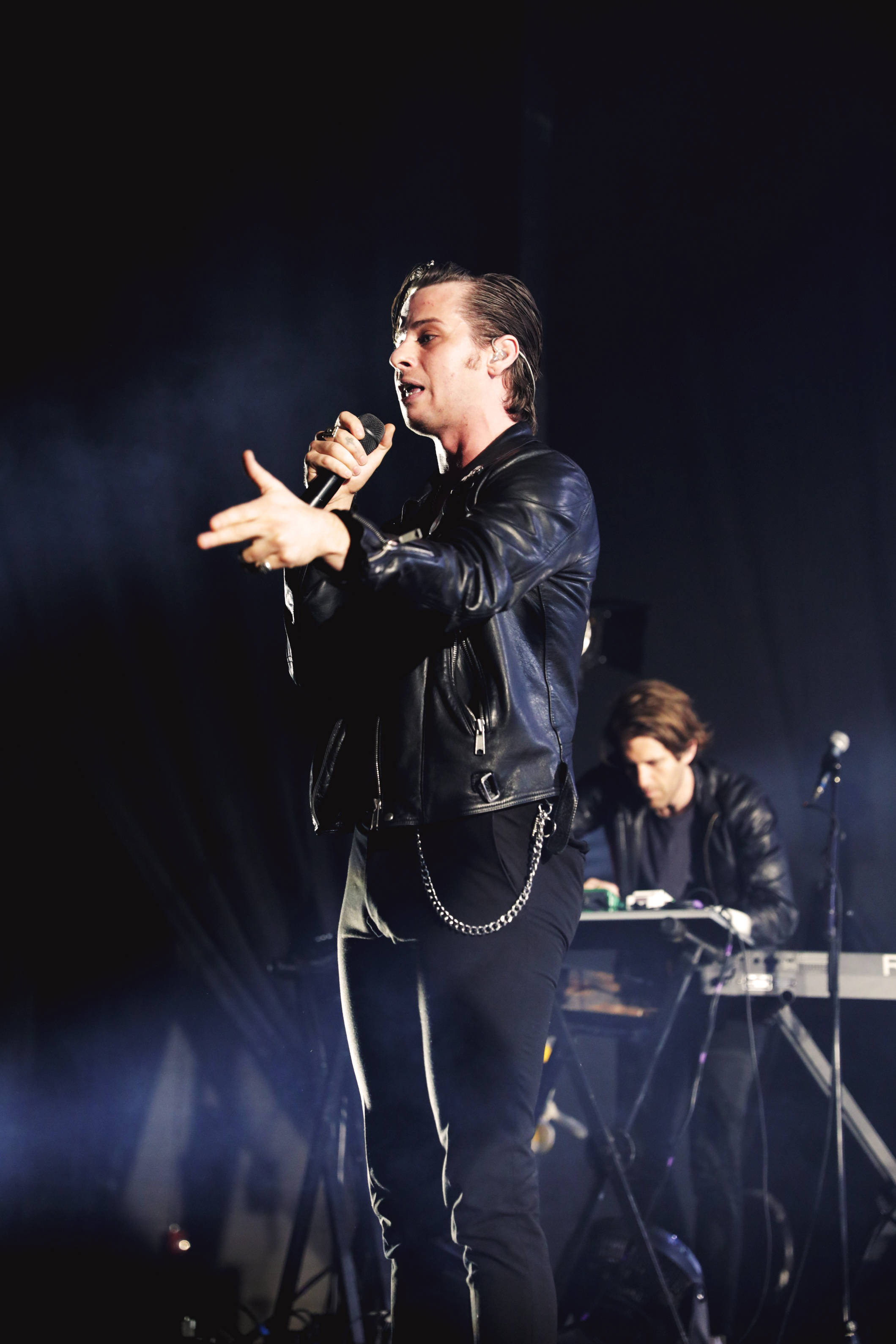 Foster the People 在黑暗舞台演出 壁紙 — 一位身穿黑色皮褸的男歌手在燈光昏暗的舞台上充滿激情地表演，背景有煙霧和一位鍵盤手。
