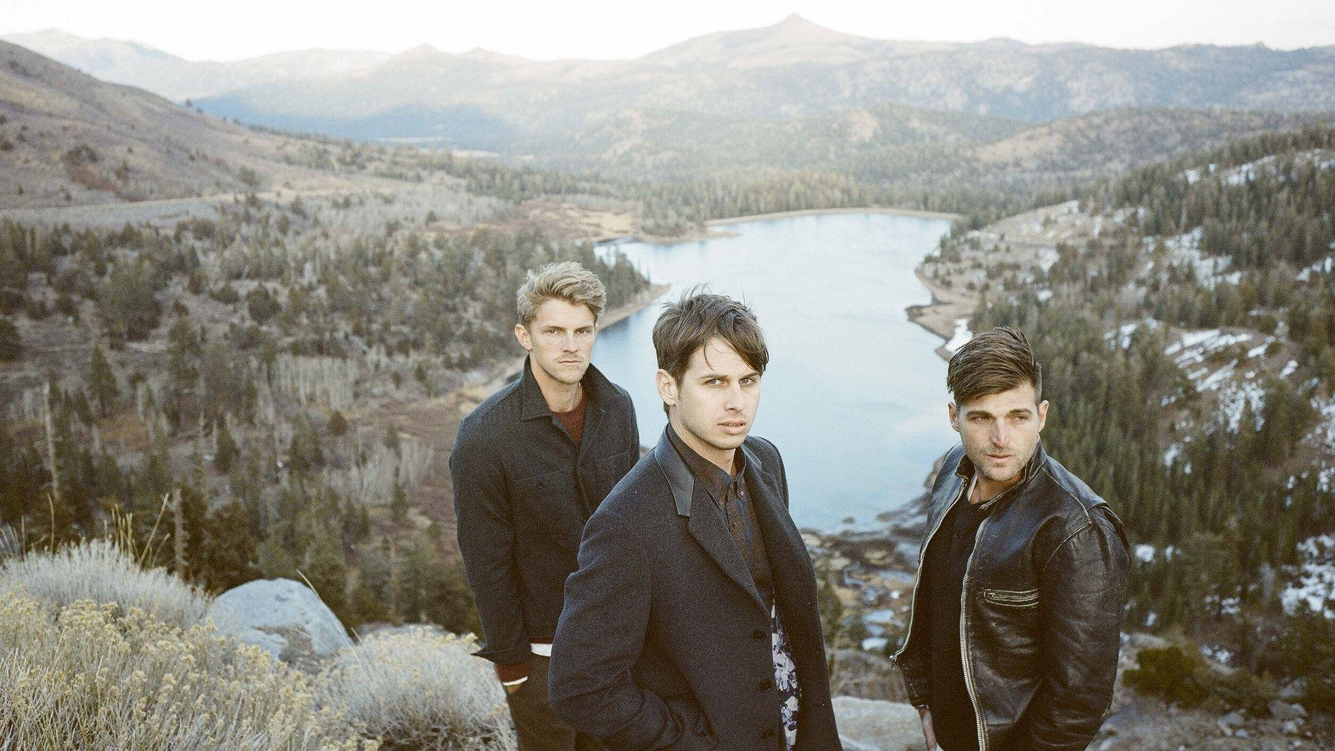 Foster The People 伴湖景 壁紙 — 三個身穿深色外套的男人站在岩石觀景點上，俯瞰著被白雪覆蓋的山脈和常綠森林環繞的蜿蜒湖泊。