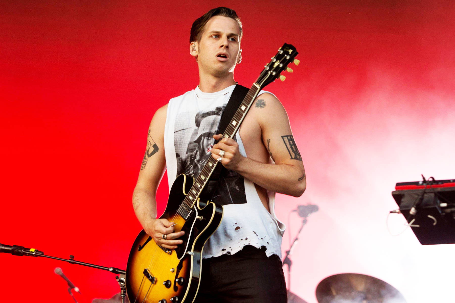 Foster The People 流行音樂樂團 壁紙 — 一位男音樂家在舞台上，以充滿煙霧效果的鮮紅色背景，彈奏一把旭日紋電結他。