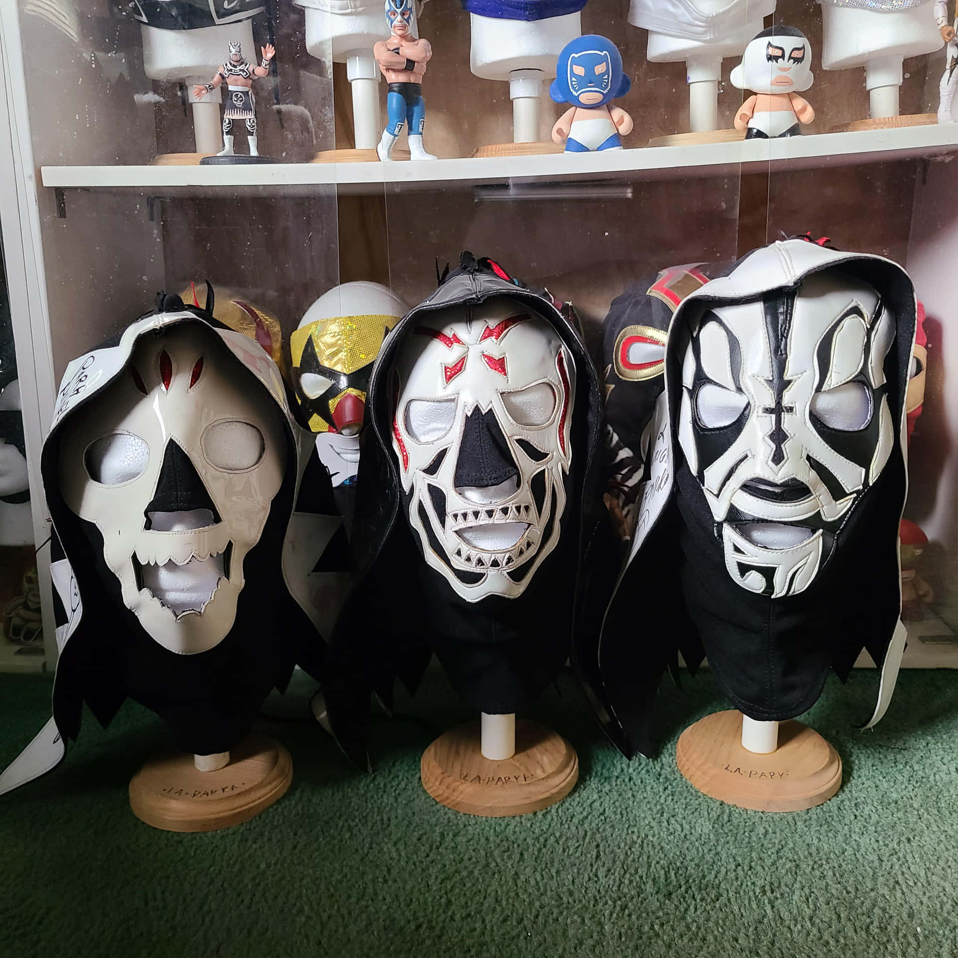  壁紙 — 四個設計精緻的墨西哥摔角面具，配有黑色頭罩，展示在木製支架上，背景為摔角人偶。