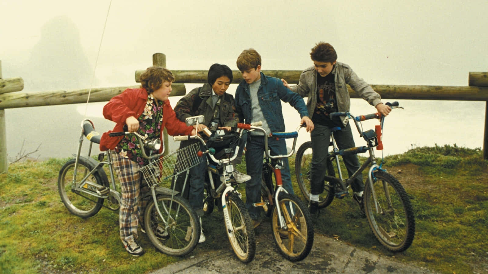  壁紙 — 四個穿著復古80年代服裝的小孩，帶著他們的BMX單車，站在霧氣瀰漫的山坡上，俯瞰水面。