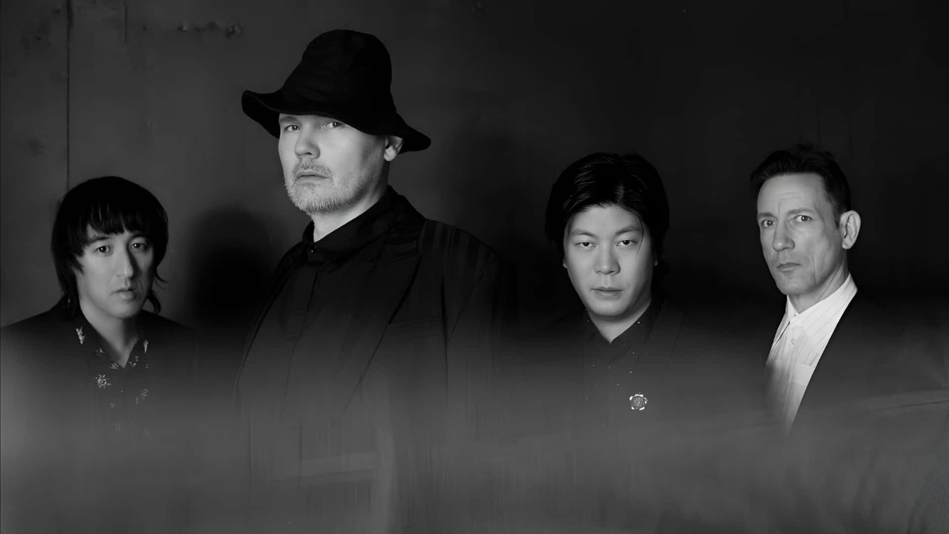  壁紙 — 四位身穿深色服裝的男子莊嚴地站在陰暗的牆前，形成一幅充滿氛圍的黑白肖像。