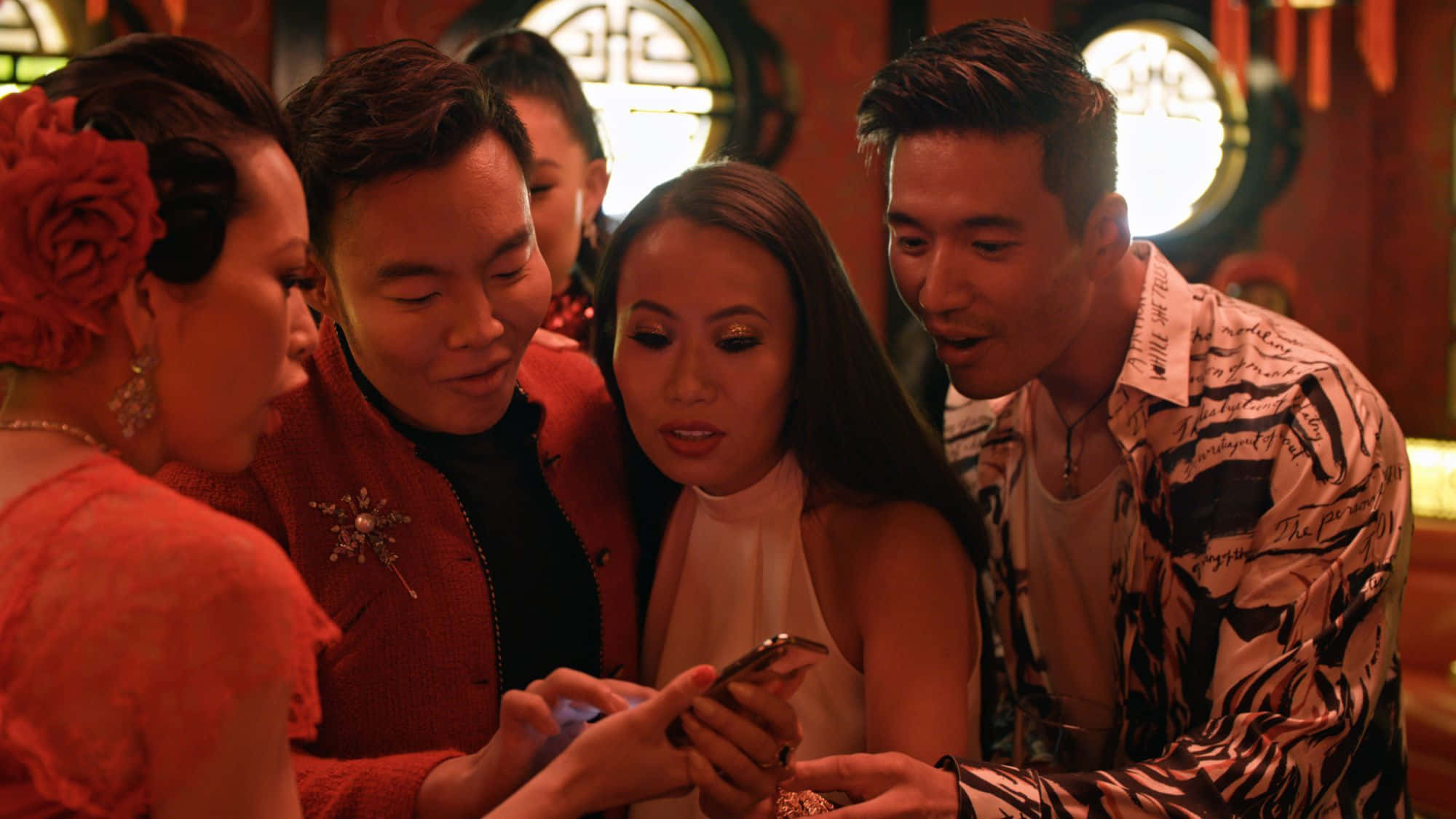 Bling Empire 桌布 壁紙 — 四個人在一個溫暖照明的房間裡緊密圍著一部智能手機，他們的表情投入且好奇。