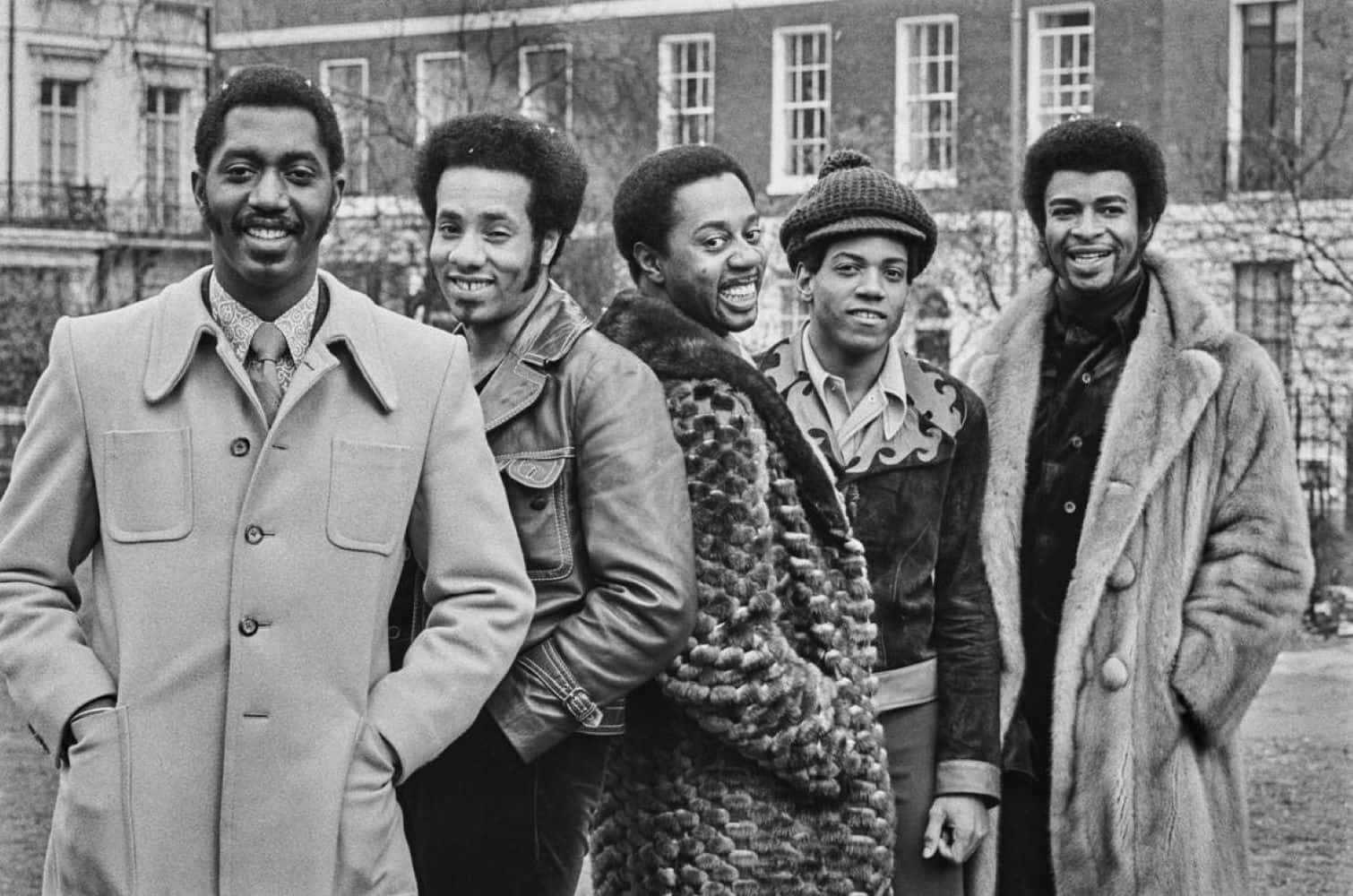 壁紙 — 四位微笑的男士穿著時尚的1970年代外套，自信地在戶外磚砌建築前擺姿勢。