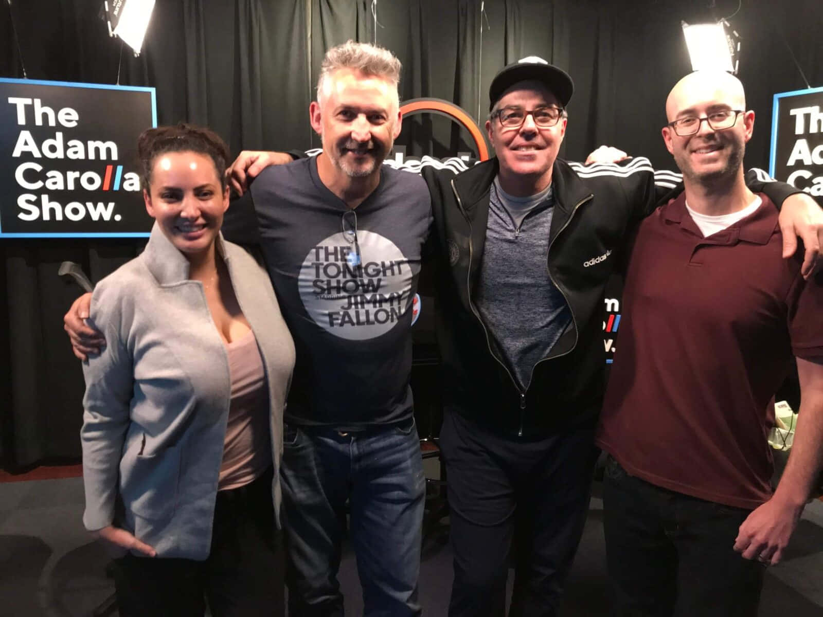  壁紙 — 四個微笑的人喺一個有「The Adam Carolla Show」背景同明亮天花燈嘅錄影廠入面一齊擺甫士。
