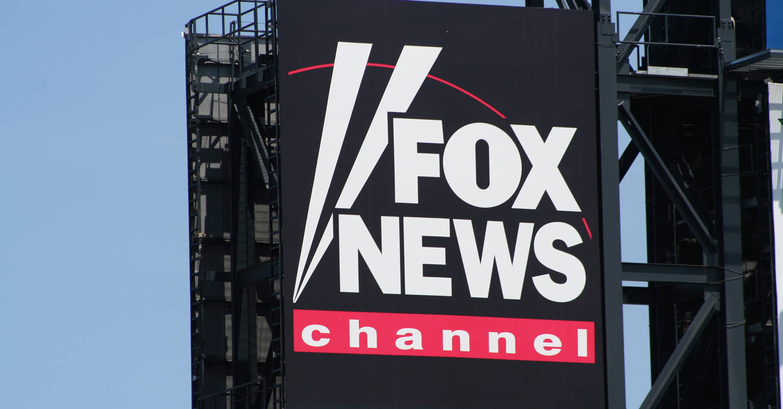 Fox News 電子廣告牌 壁紙 — 一個大型黑色Fox News Channel標誌，帶有白色和紅色字體，安裝在金屬結構上，背景是清澈的藍天。