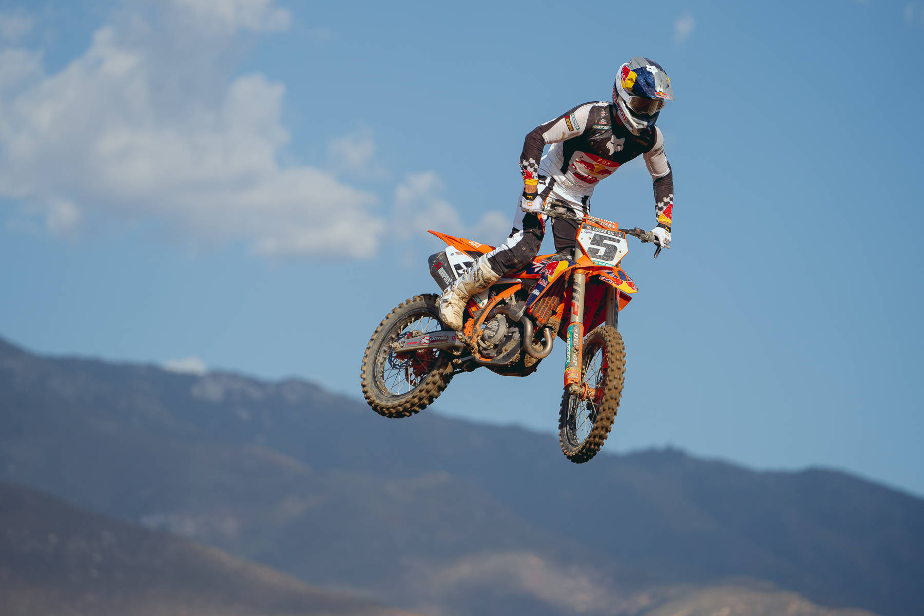 Fox Racing 沙灘摩托車高跳特技 壁紙 — 一位越野電單車手騎著橙色電單車在空中飛翔，背景是遠山和晴朗的藍天。