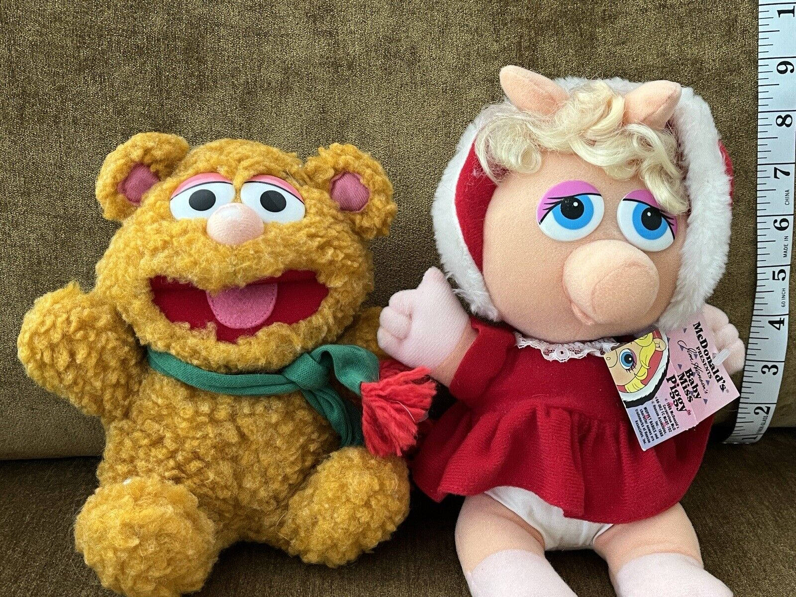 Fozzie Bear 與 Miss Piggy 壁紙 — 兩個毛絨Muppet公仔，一隻戴綠色圍巾的黃色熊和一隻穿著紅色冬季服裝的豬，並排坐在棕色沙發上，旁邊有一個軟尺。