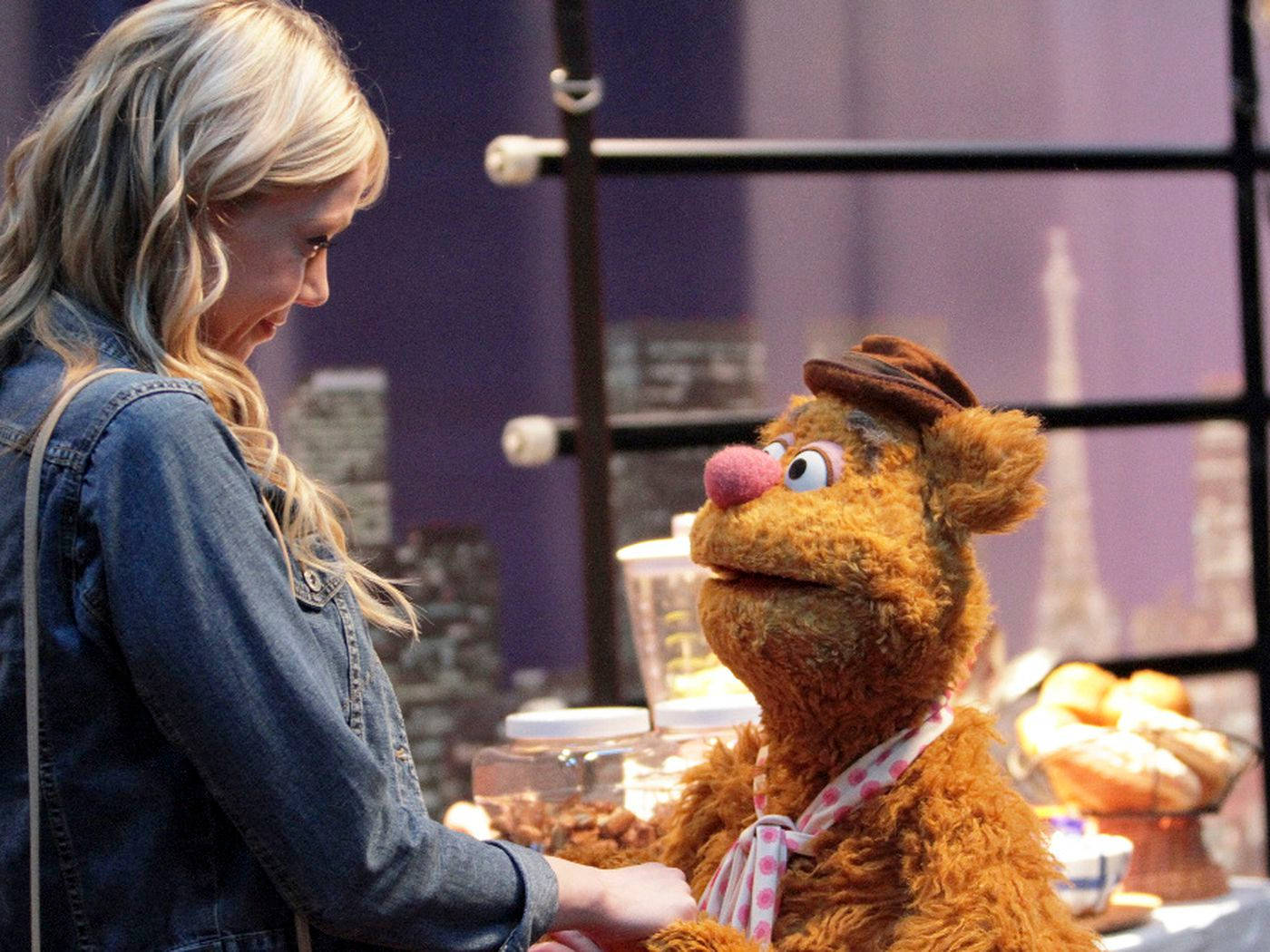 Fozzie Bear 與 Olivia Wilde 壁紙 — 一位金髮女子穿著牛仔外套，在麵包店櫃檯與一隻戴著帽子和圍巾的毛茸茸棕色木偶熱情互動。