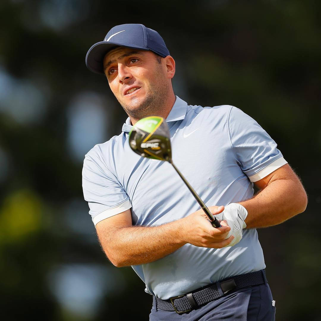 Francesco Molinari 高爾夫揮杆 壁紙 — 一位專注的男性高爾夫球手正在揮桿，戴著藍色帽子，身穿淺藍色Polo衫，背景是模糊的綠色。
