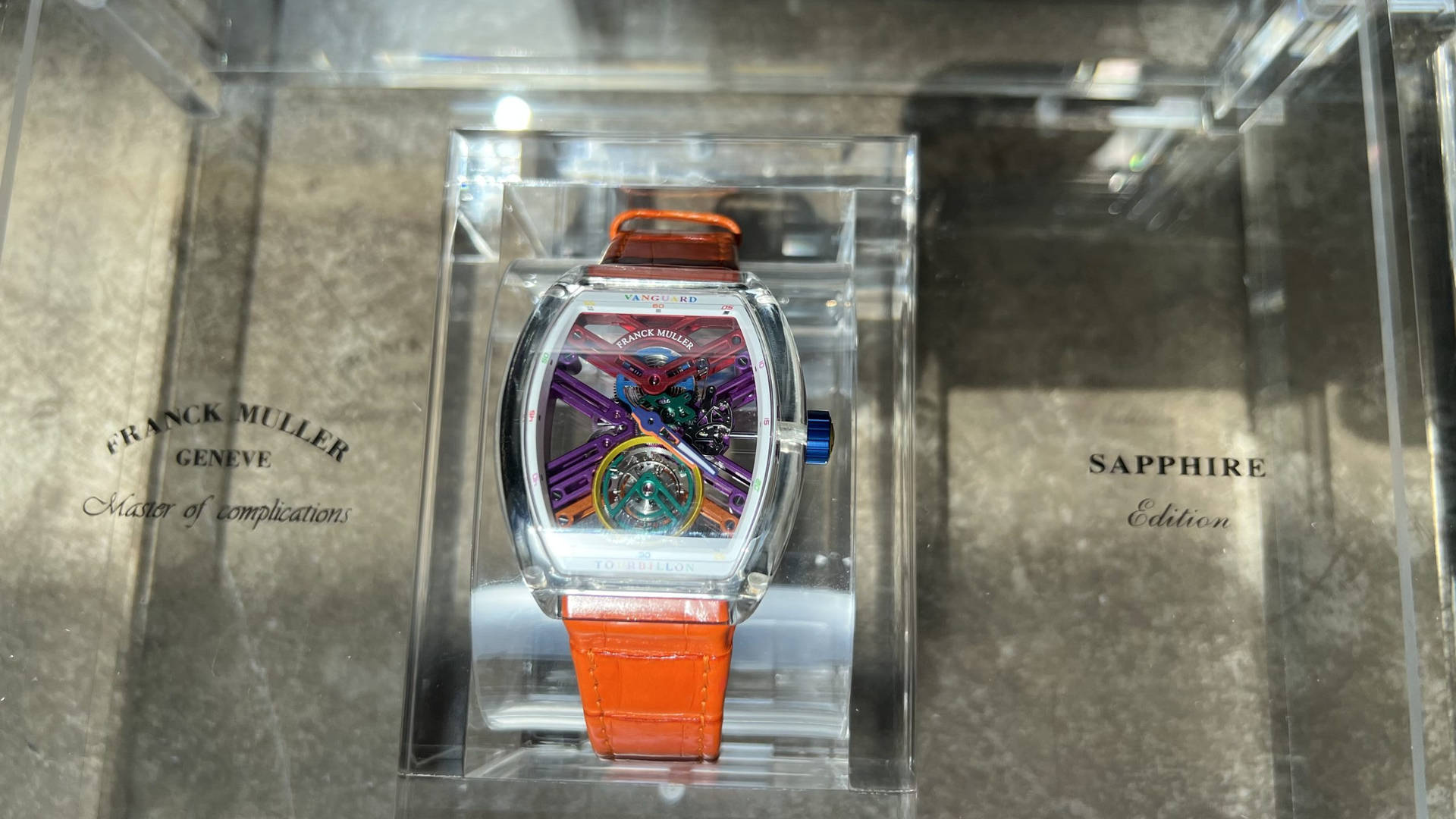 Franck Muller 奢華手錶配橙色錶帶，置於藍寶石水晶錶盒中，在大理石展示台上。
