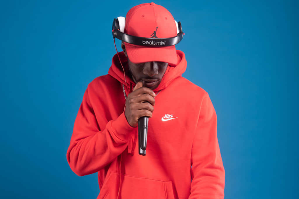 深入創意世界：Frank Ocean筆電 壁紙 — 一位穿著紅色Nike連帽衫和Jordan帽子的說唱歌手，在純藍色工作室背景前對著麥克風說唱。
