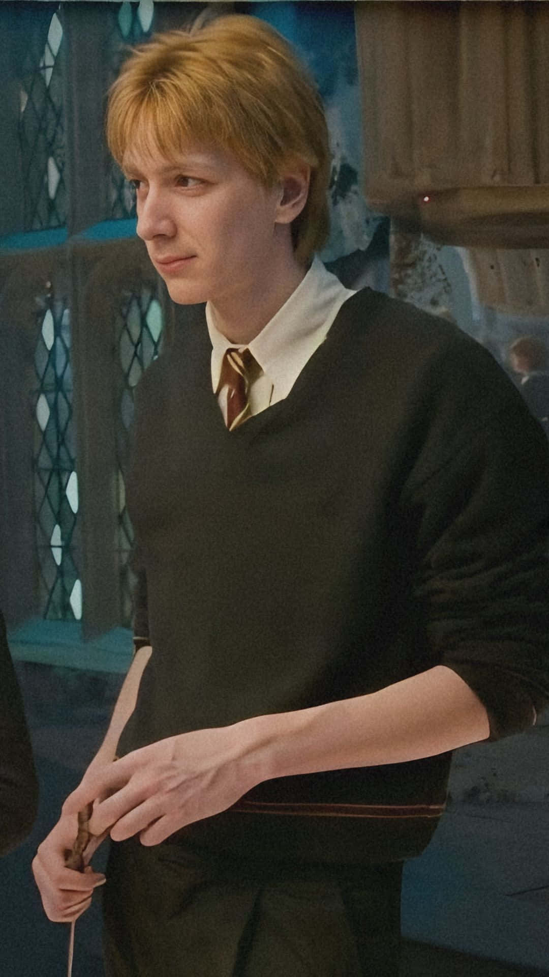 Fred Weasley 的休閒霍格華茲造型 壁紙 — 一位金髮青年穿著深色毛衣和條紋領帶，在一個光線昏暗、有彩繪玻璃窗的石室中手持魔杖。