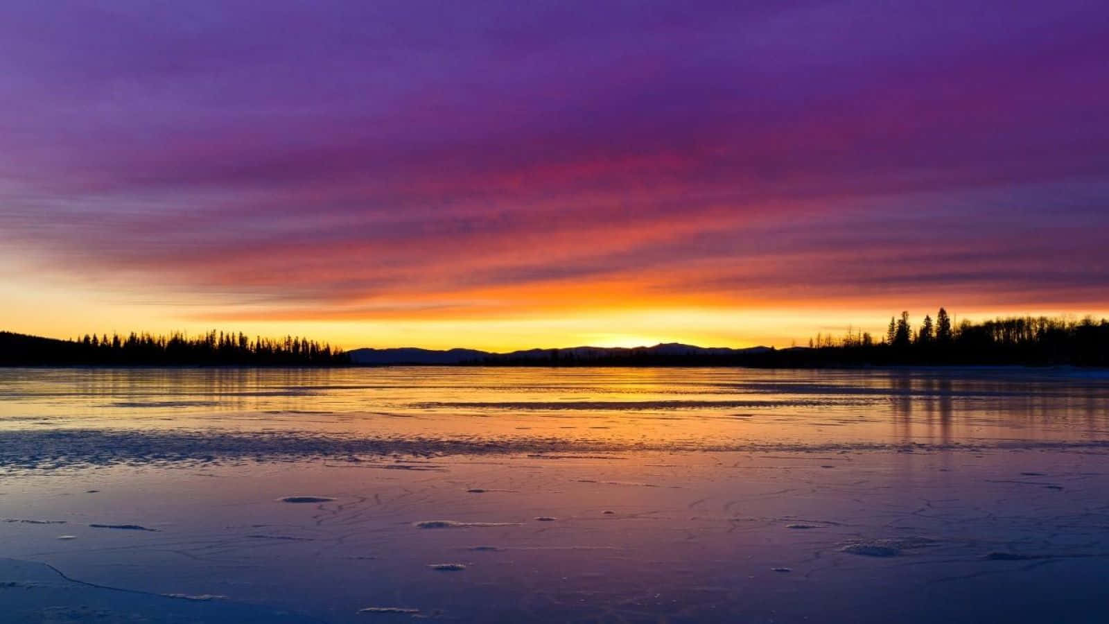 Sunset Reflection 桌布 壁紙 — 結冰的湖面映照出夕陽的火焰色彩，紫色、橙色和黃色在遠處樹木覆蓋的山丘上閃耀。
