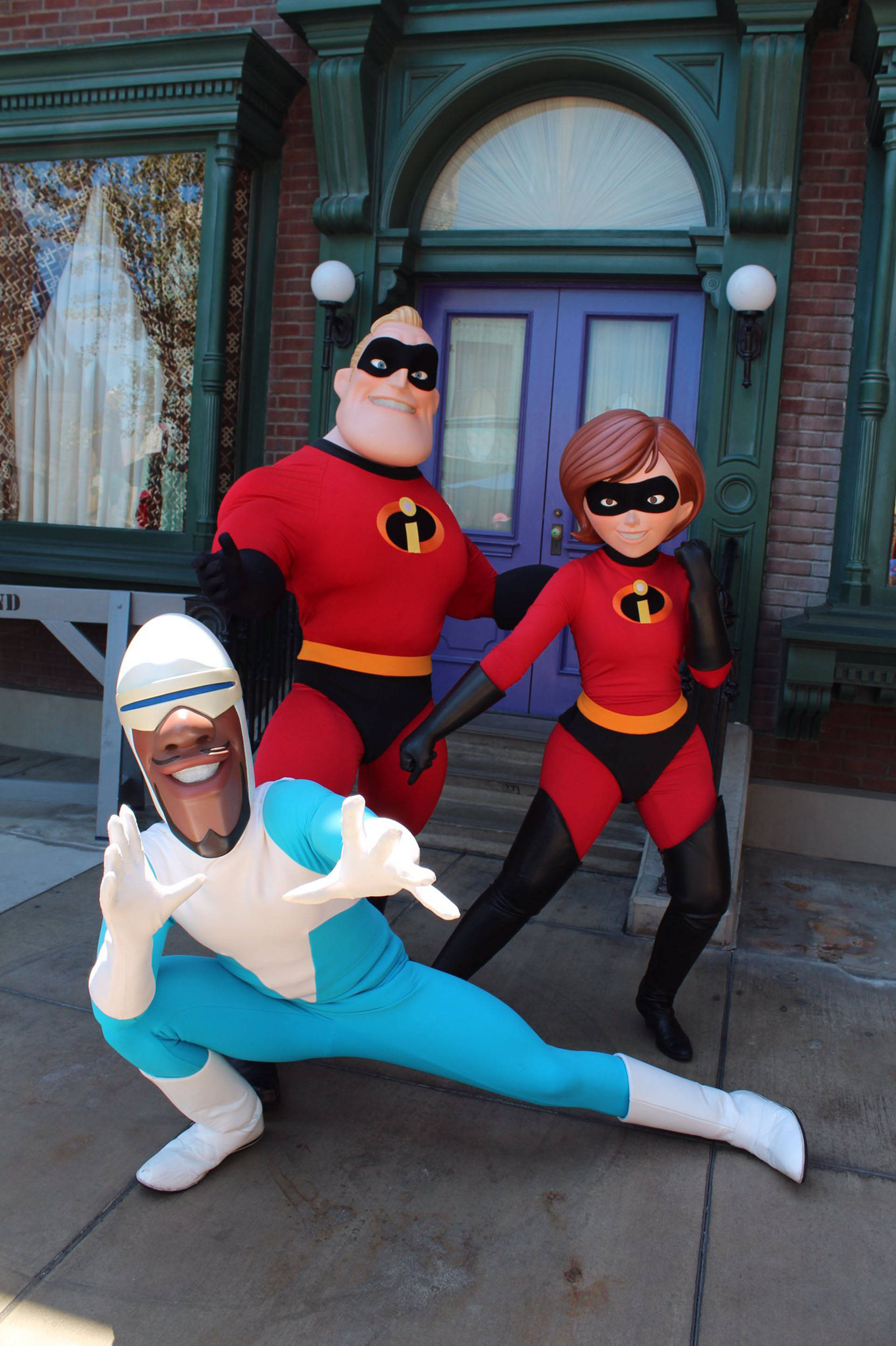 Frozone 優雅姿勢 壁紙 — 三個身穿全套服裝的超人特攻隊角色在一個有綠色飾邊和紫色門的主題建築外愉快地擺姿勢。