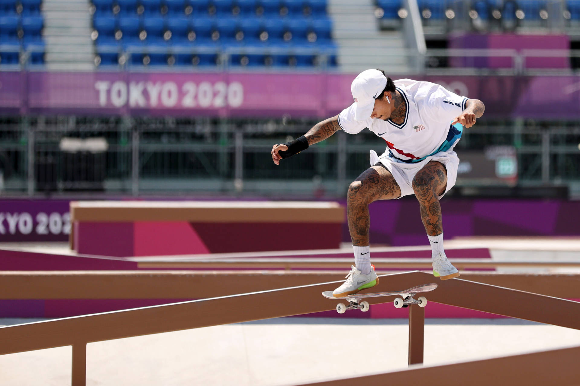 FS Feeble 滑板動作 壁紙 — 一位紋身滑板手穿著白色裝備，在明亮的日光下於東京2020奧運會上磨軌。