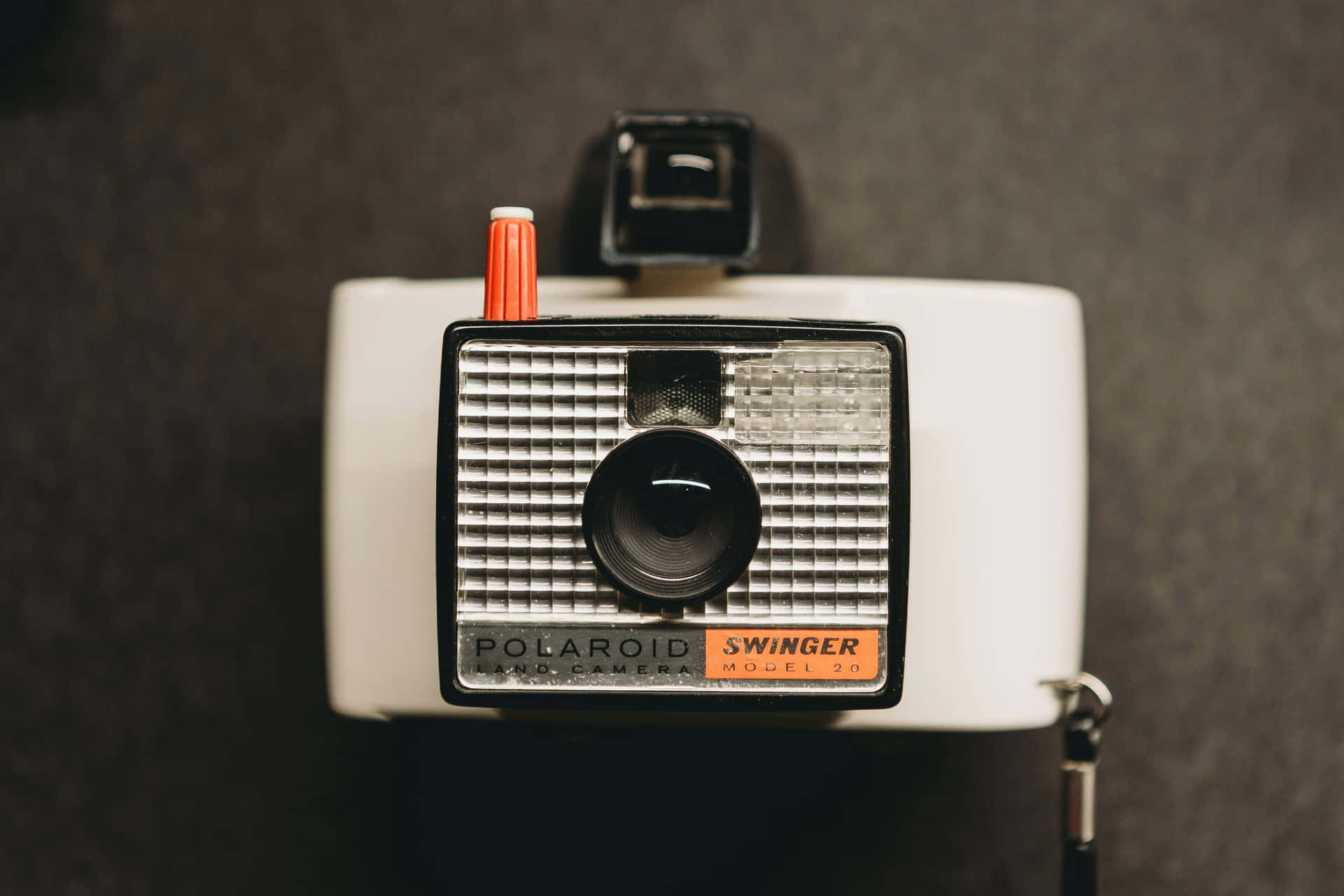  壁紙 — 一部復古白色Polaroid Swinger Land相機，帶有橙色快門按鈕，靠在深色紋理牆上。