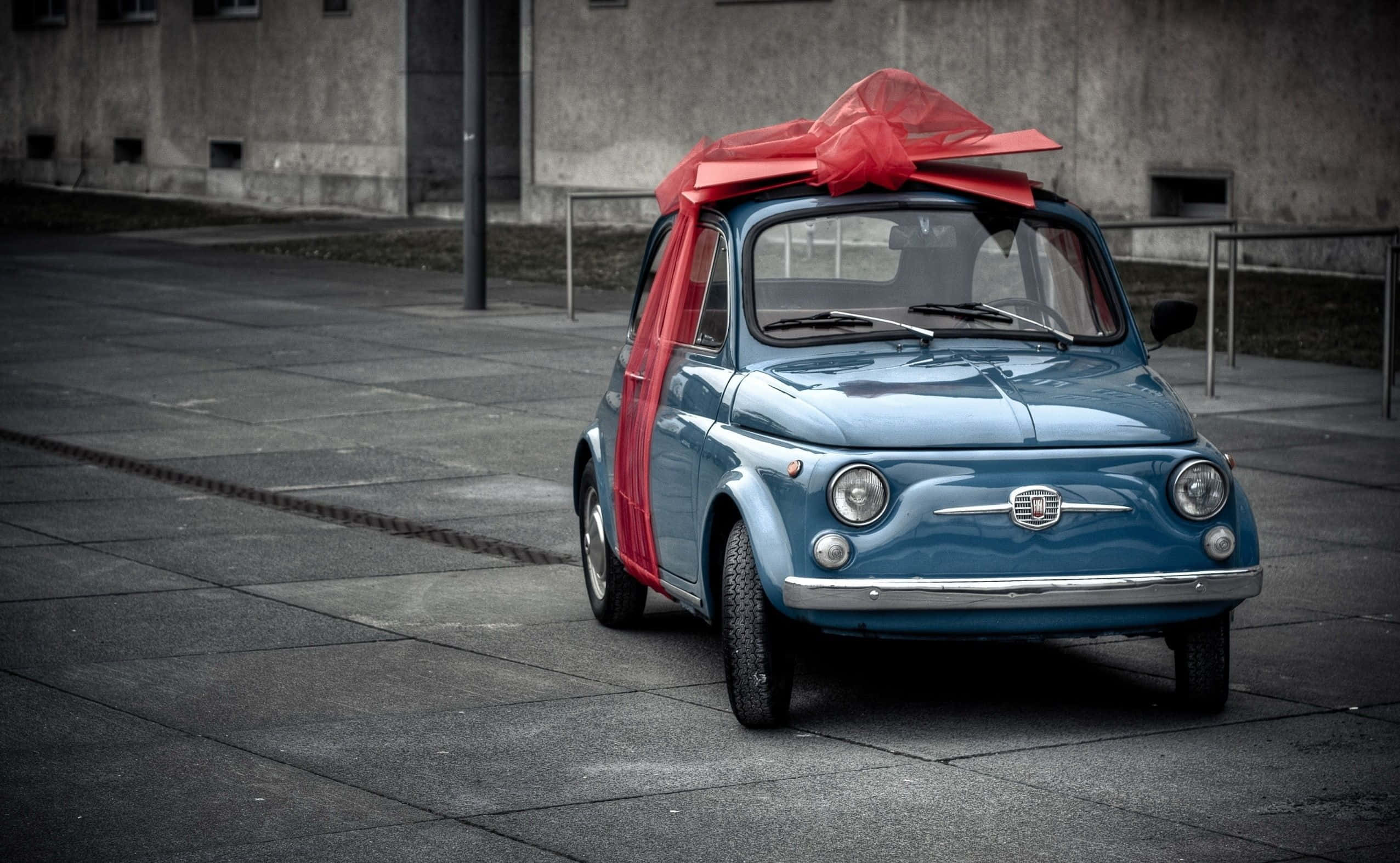 Fiat 500l 桌布 壁紙 — 一輛復古藍色菲亞特500，飾有一個大紅色蝴蝶結，停在灰色混凝土廣場上，背景是風化的工業建築。