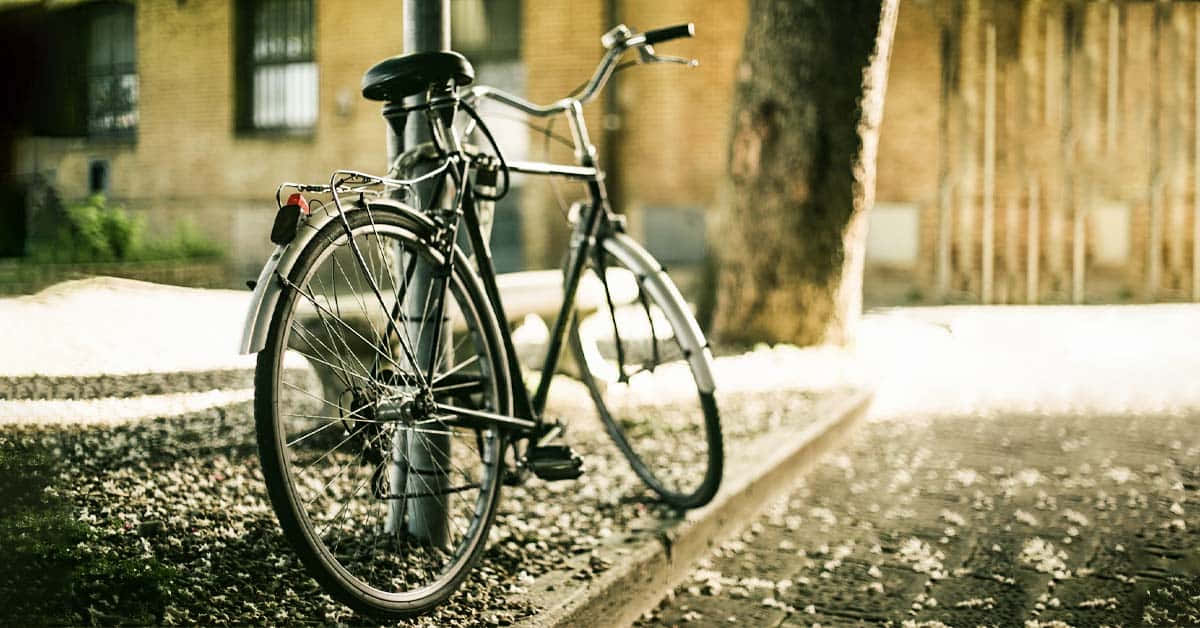  壁紙 — 一輛復古單車靠在一根柱子上，旁邊是陽光普照的人行道、一棵樹和磚砌建築。