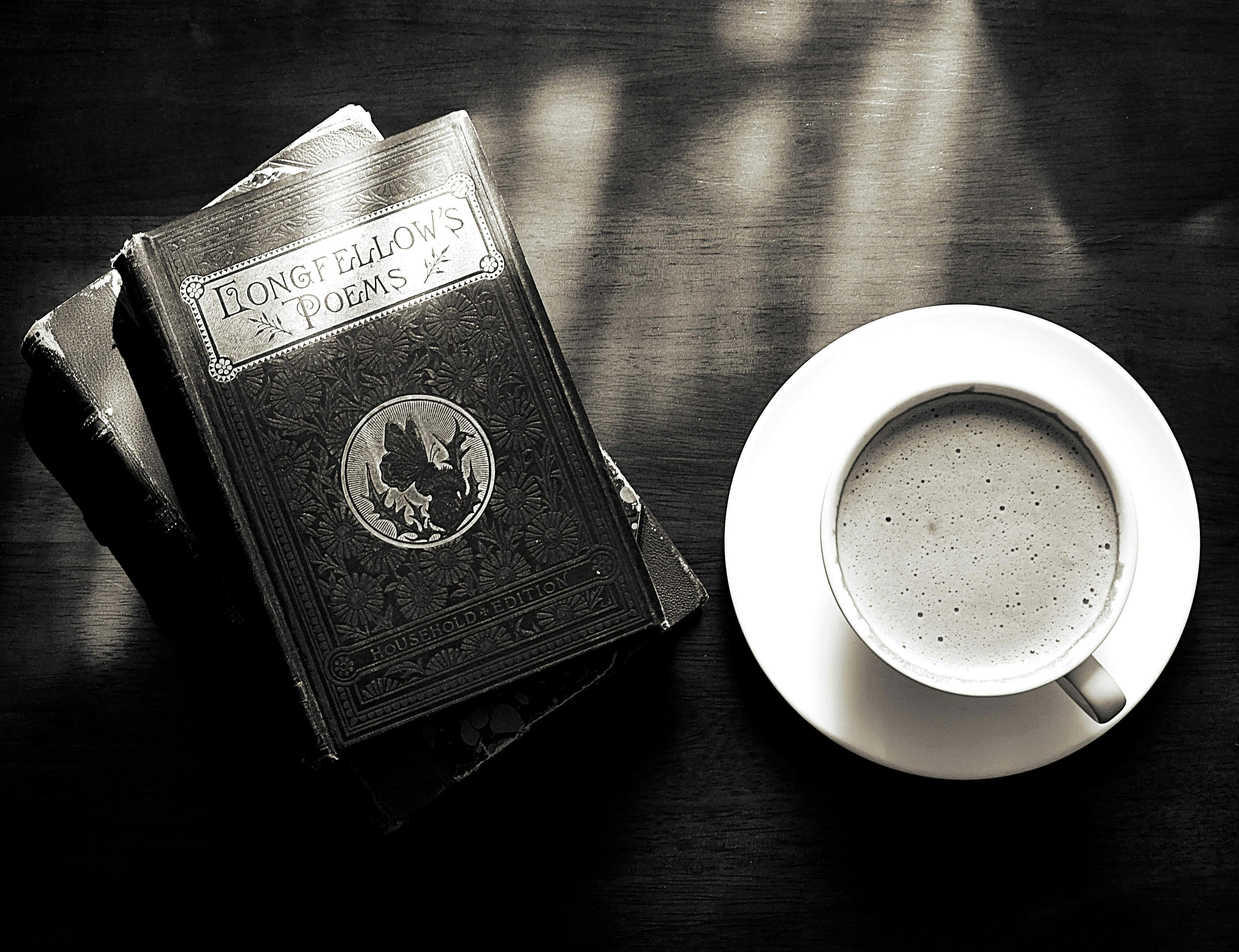 Dark Book Aesthetic 桌布 壁紙 — 一本復古的朗費羅詩集放在一張深色木桌上，旁邊是一杯冒著熱氣的咖啡，沐浴在柔和的陽光中。