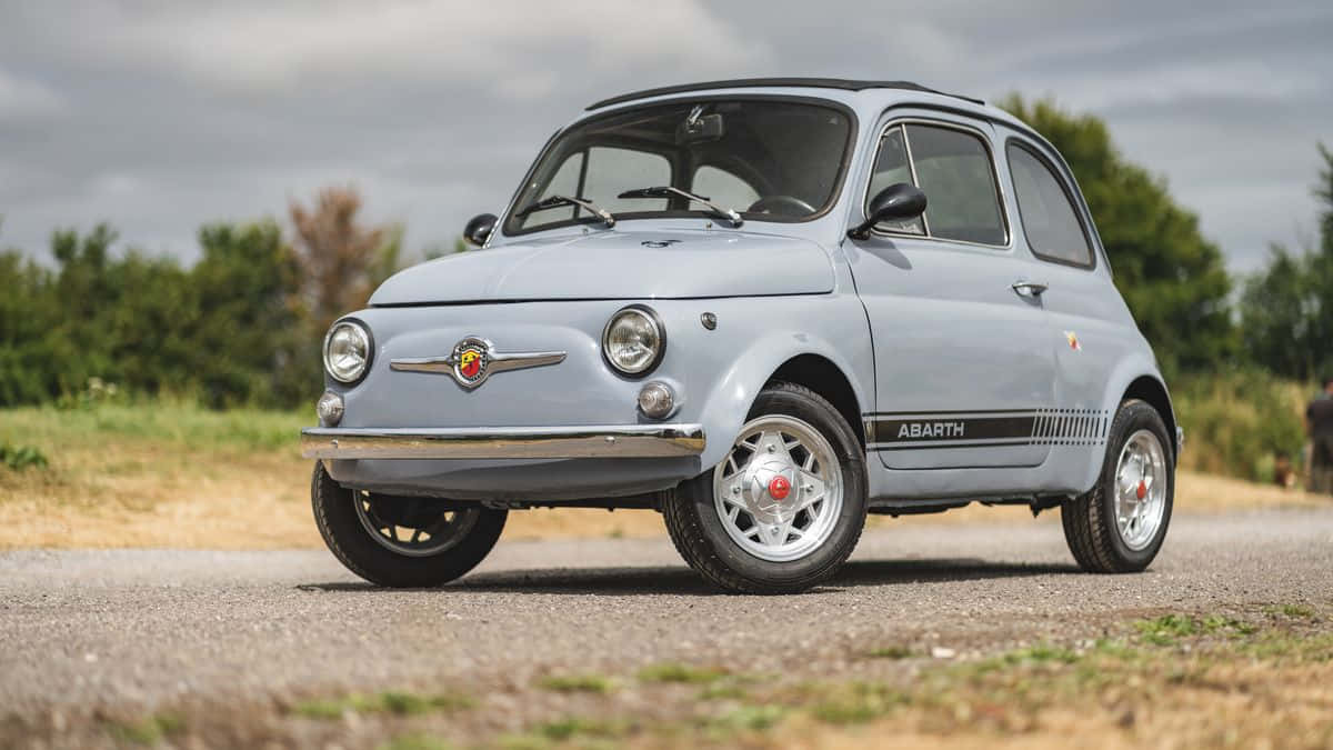 Fiat 500 桌布 壁紙 — 一架復古淺藍色Fiat 500 Abarth停泊在碎石路上，背景有綠樹，天空多雲。