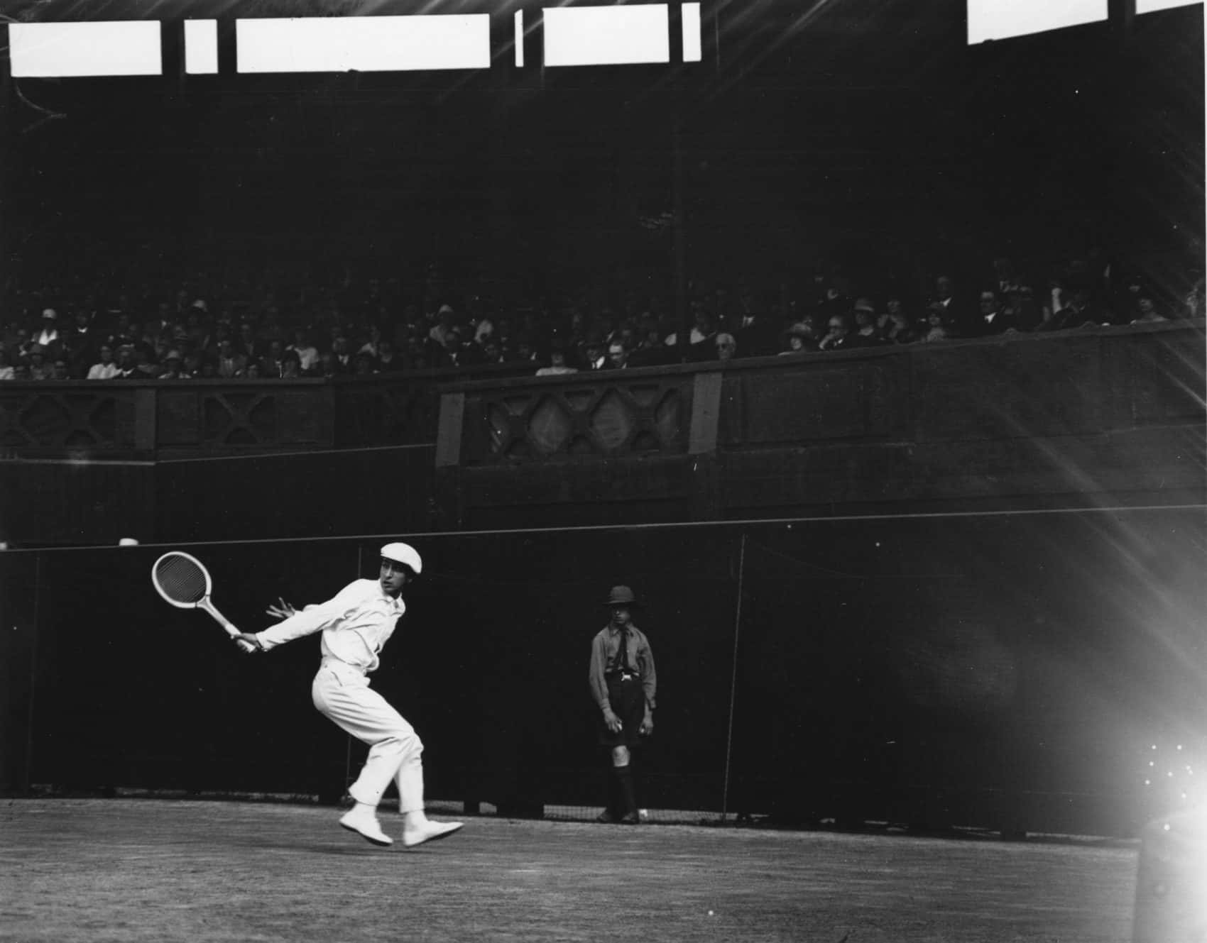 René Lacoste 擊球瞬間 壁紙 — 一張復古黑白照片捕捉到一位網球運動員在擠滿觀眾的室內場館中揮拍，燈光效果戲劇性。