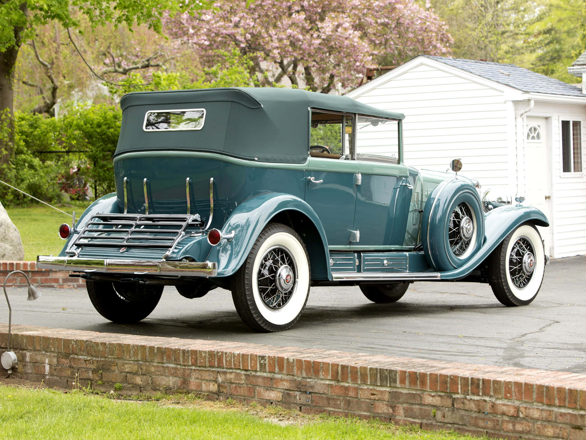 Cadillac Fleetwood 桌布 壁紙 — 一輛復古藍綠色敞篷車配白色輪胎，停在磚砌邊緣上，旁邊是白色棚屋，上方有盛開的樹木。