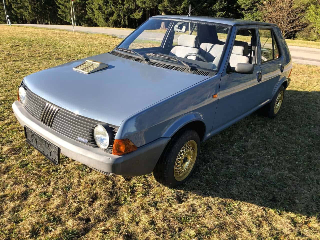 Fiat Ritmo 桌布 壁紙 — 一輛復古藍色快意烏諾停泊在乾草地上，背景有樹木，在明亮的陽光下。