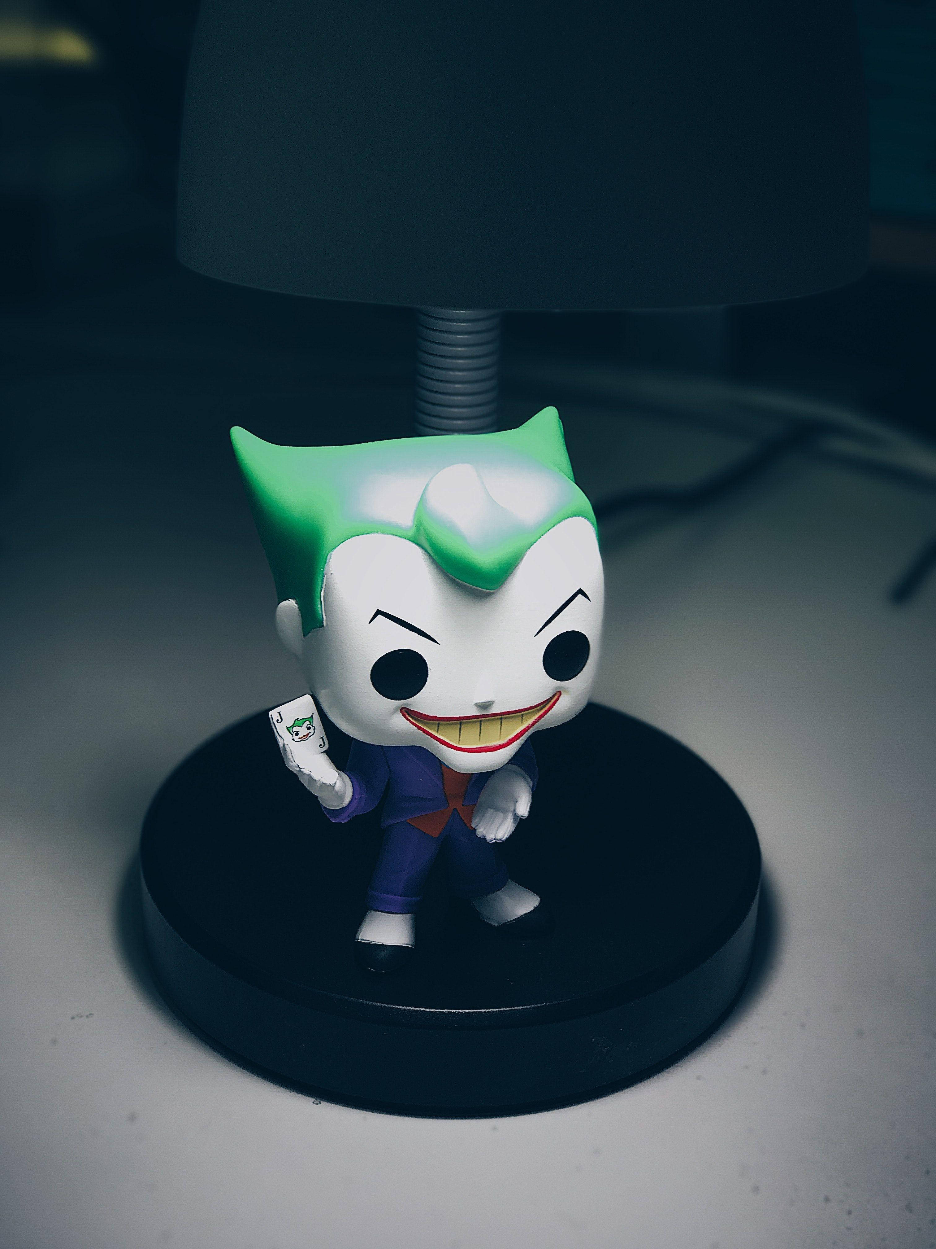 Funko Pop 小丑形象 壁紙 — 一個Funko Pop! 小丑公仔，綠色頭髮，紫色西裝，在白色表面上，暗燈下拿著一張小丑牌。