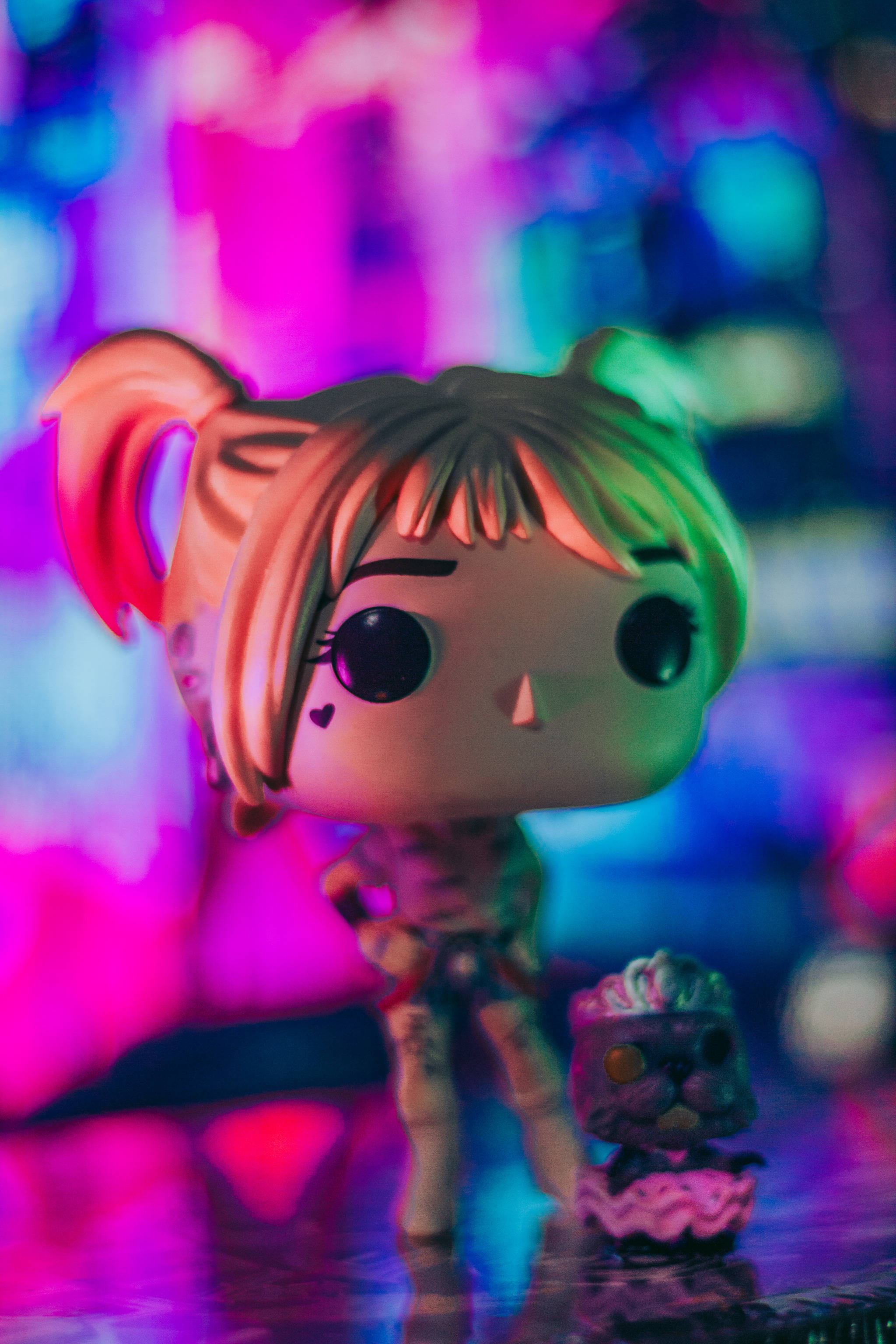 Funko Pop 哈莉·奎茵 壁紙 — 一個紮辮子的Funko Pop公仔站在一個小蛋糕旁邊，在充滿活力的粉紅色和藍色霓虹燈下。