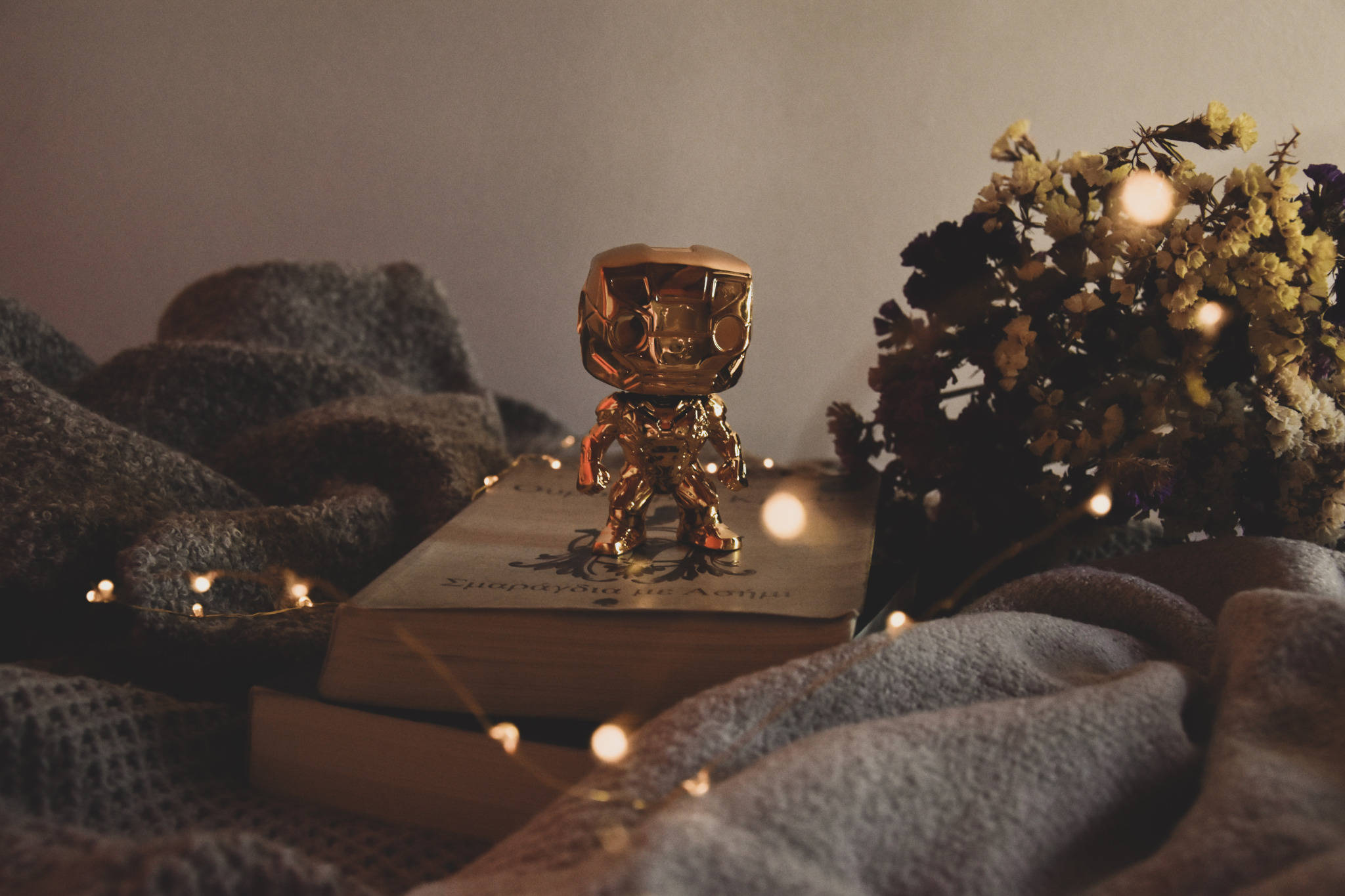 Funko Pop 鋼鐵人金屬版 壁紙 — 一個金色機械人公仔企喺一本書上面，周圍有舒適嘅毛氈同閃爍嘅仙女燈。