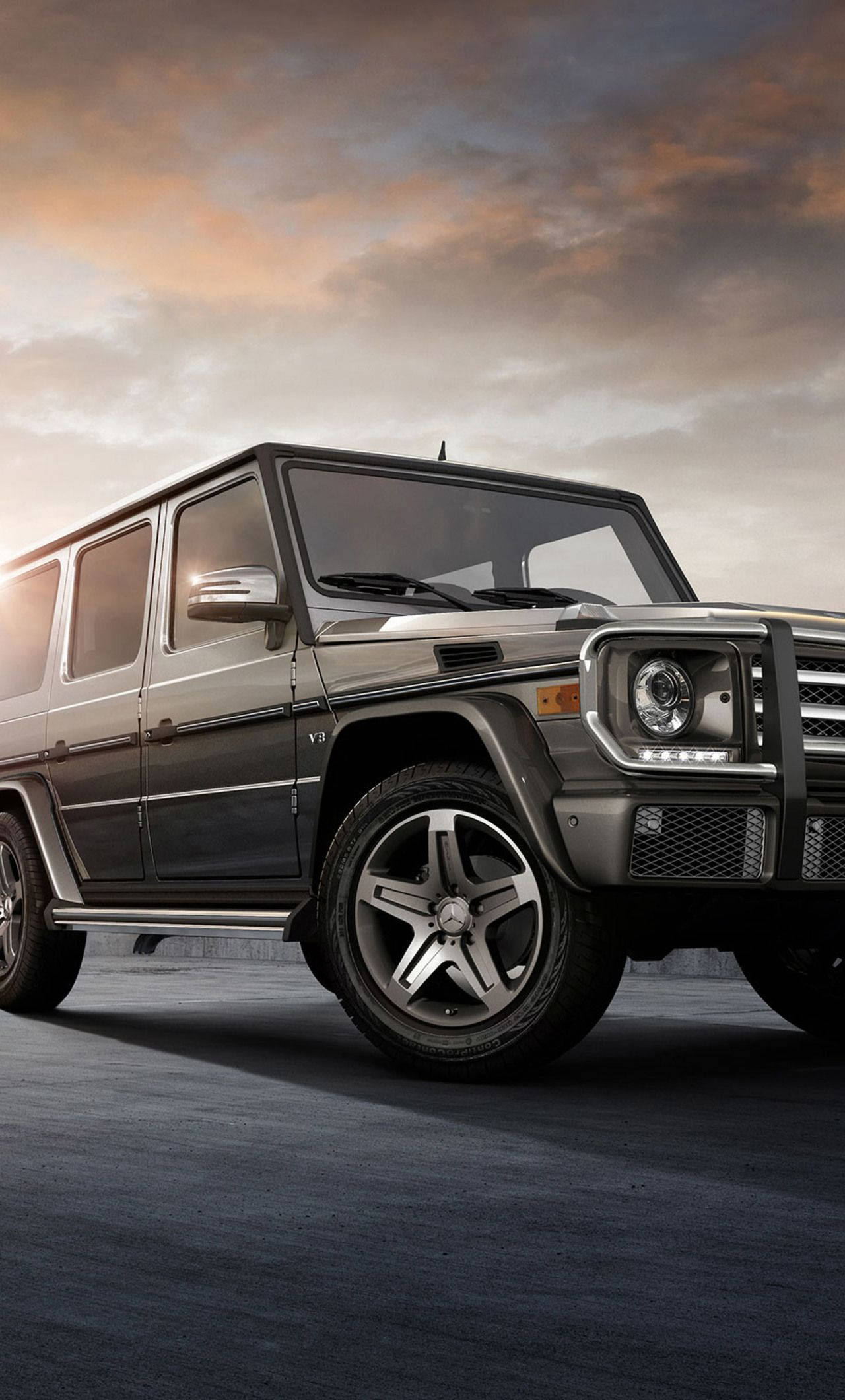 梅賽德斯-奔馳 G-Class iPhone 版 壁紙 — 一輛時尚的深灰色梅賽德斯 G-Class SUV 停在混凝土表面上，背景是戲劇性的日落天空，帶有溫暖的橙色和灰色雲彩。