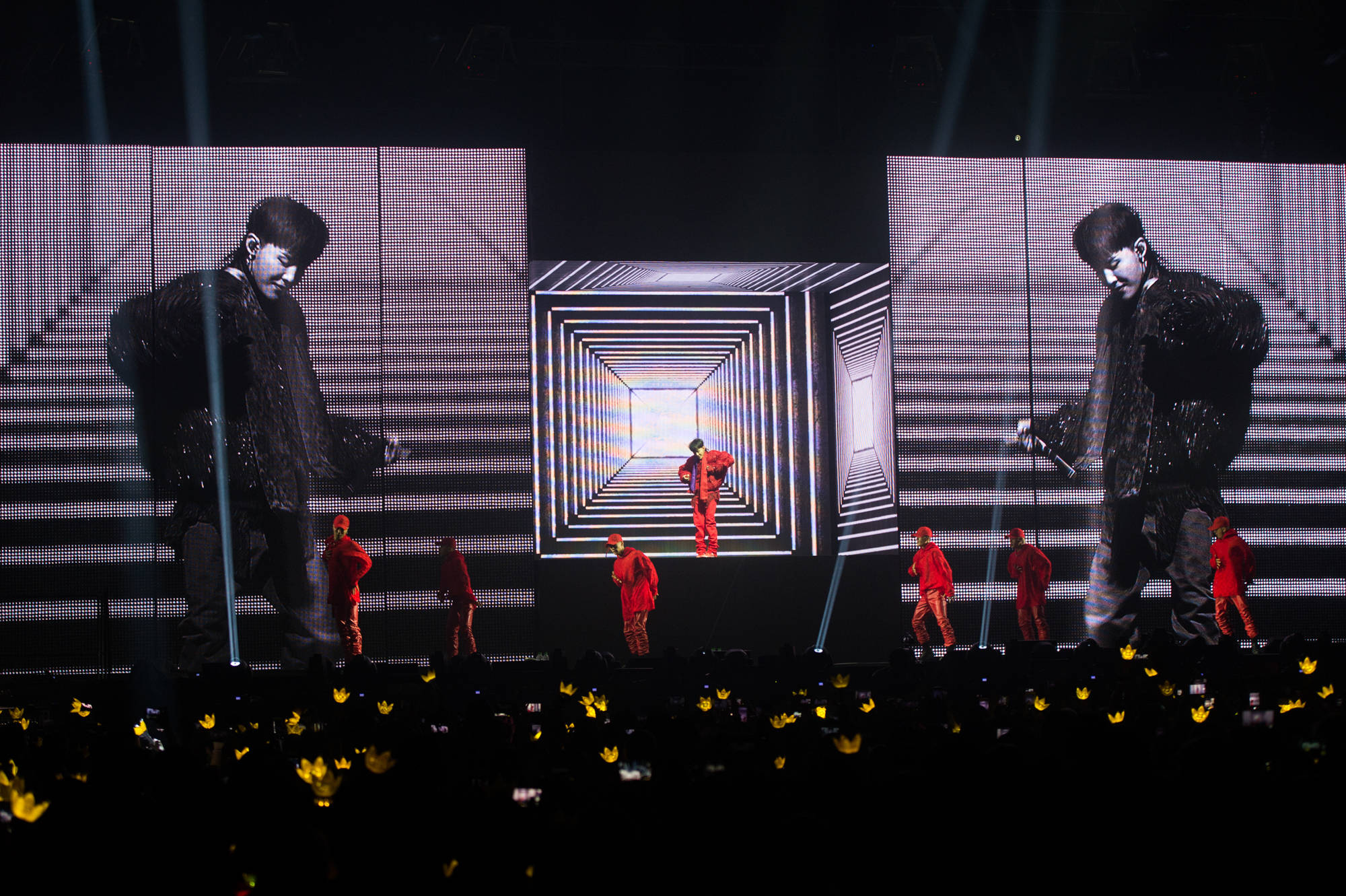 G-dragon MOTTE演唱會 壁紙 — 一位身穿紅色表演者站在舞台中央，處於發光的數碼隧道內，兩旁的大屏幕顯示他的剪影，紅色舞者在他周圍移動，聚光燈下，觀眾在下方舉著黃色燈光。