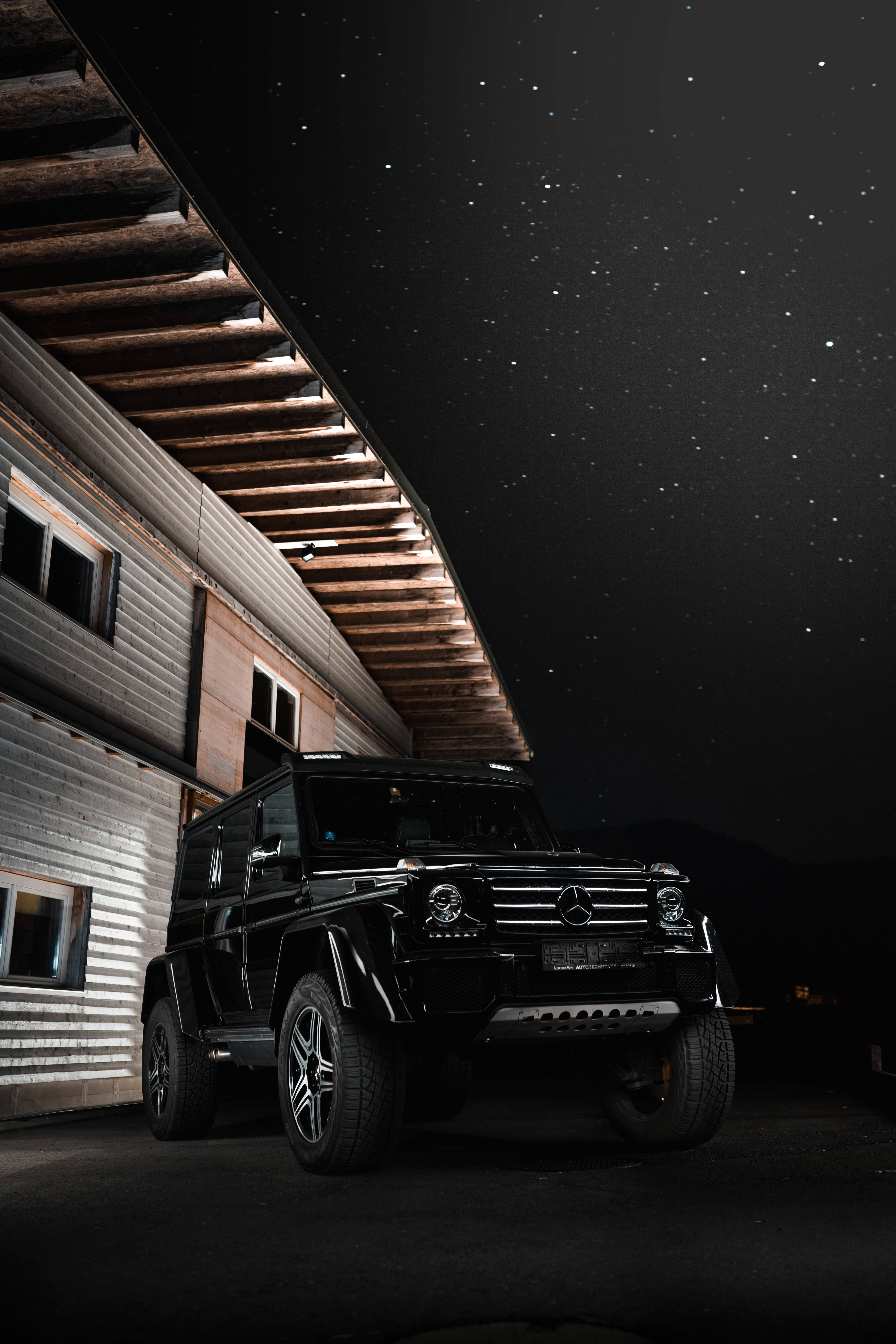夜間梅賽德斯-奔馳G-Wagen 壁紙 — 一輛黑色梅賽德斯G-Wagen停泊在星空夜空下，旁邊是一座帶有照明木樑的現代建築。