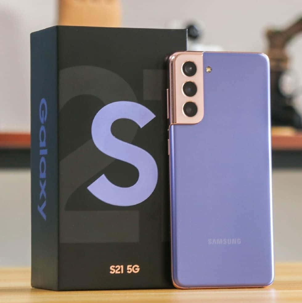 以革命性Galaxy S10保持連線 壁紙 — 一部時尚的薰衣草色Samsung Galaxy S21 5G智能手機站在木質表面上，旁邊是它的黑色盒子。