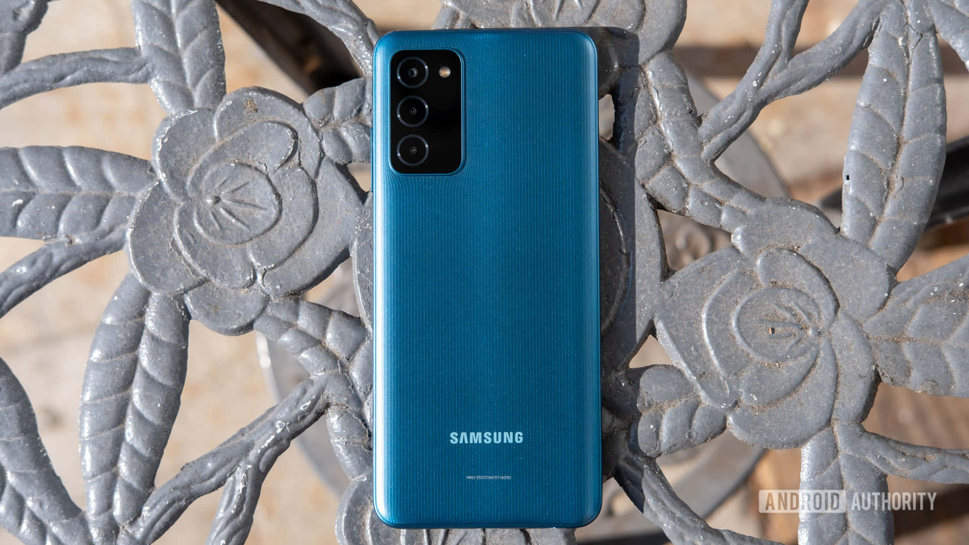 三星 Galaxy S21 Ultra 壁紙 — 一部充滿活力的藍色Samsung智能手機直立放在一張帶有花卉圖案的華麗灰色金屬長椅上。