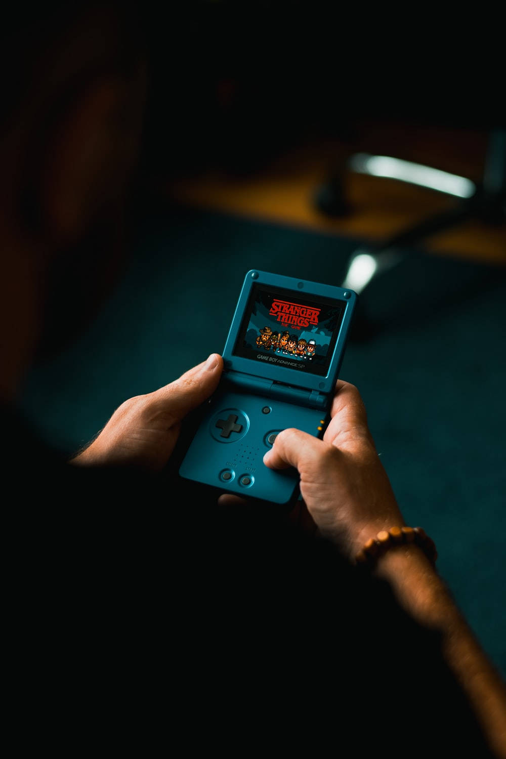 Game Boy Advance SP《怪奇物語》 壁紙 — 一個人在燈光昏暗的房間裡手持一部顯示《怪奇物語》遊戲的藍色手提遊戲機。